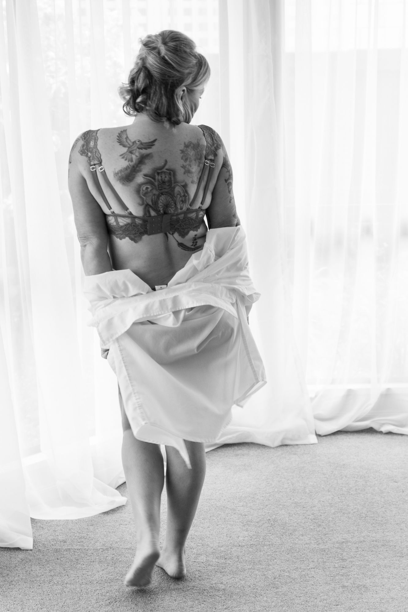Black and white rear view of tattooed woman in lace bra removing white shirt near window. Boudoir photography Christchurch