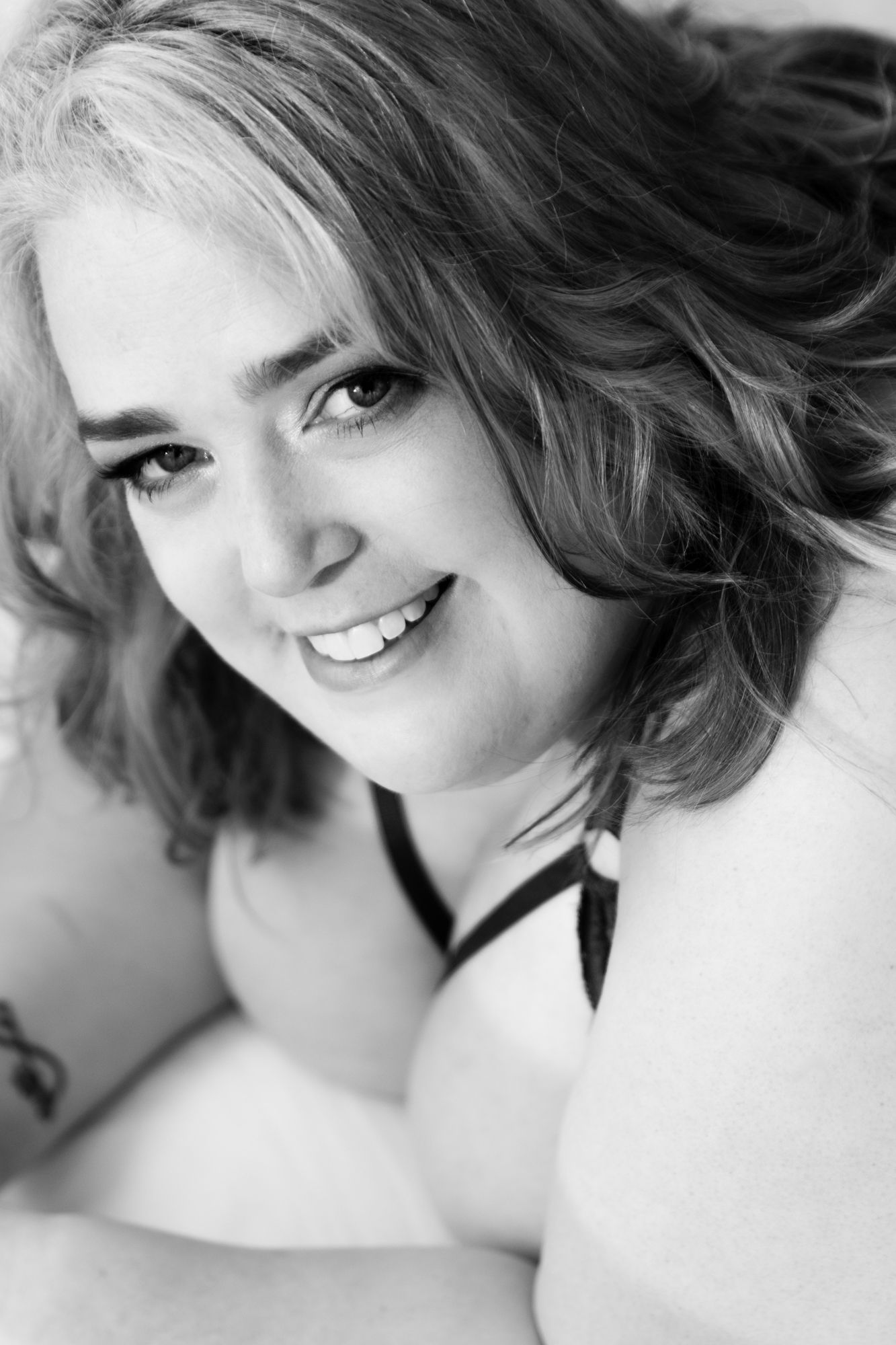 Black and white close-up portrait of a smiling woman with wavy hair wearing a dark bra strap, lying down. Boudoir photography Christchurch