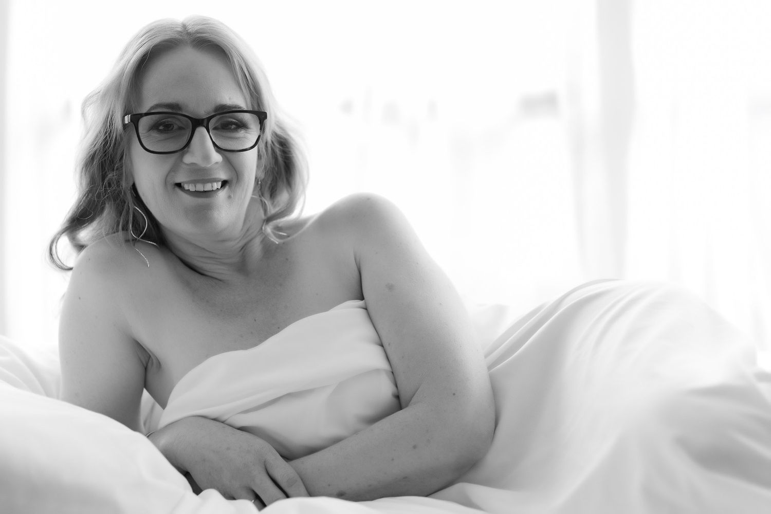 Black and white boudoir photo of smiling woman with glasses wrapped in white sheet on bed. Boudoir photography Christchurch