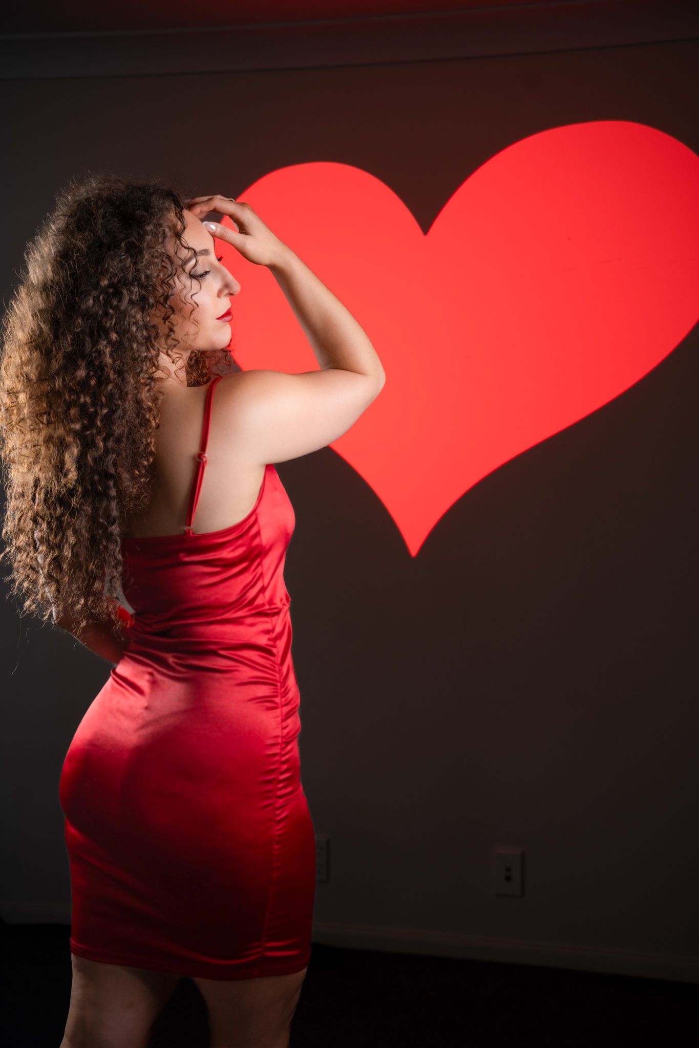 Woman in red satin dress poses with hand raised before a large red heart projection on dark wall. Boudoir photography Christchurch portrait