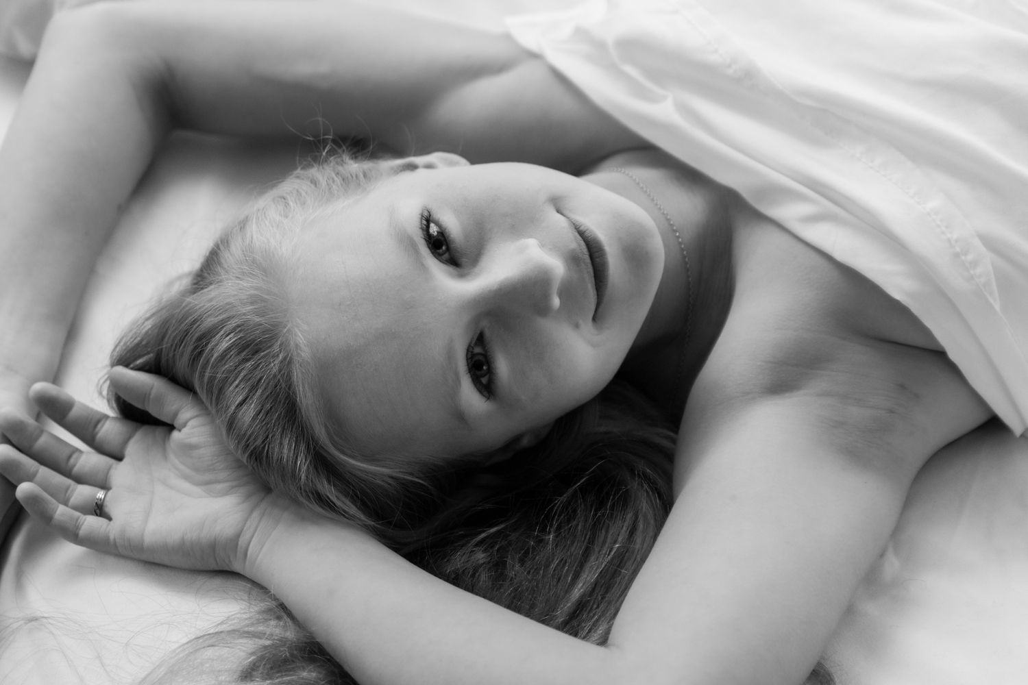 Close-up black and white portrait of smiling woman lying on white bedding looking at camera. Boudoir photography Christchurch