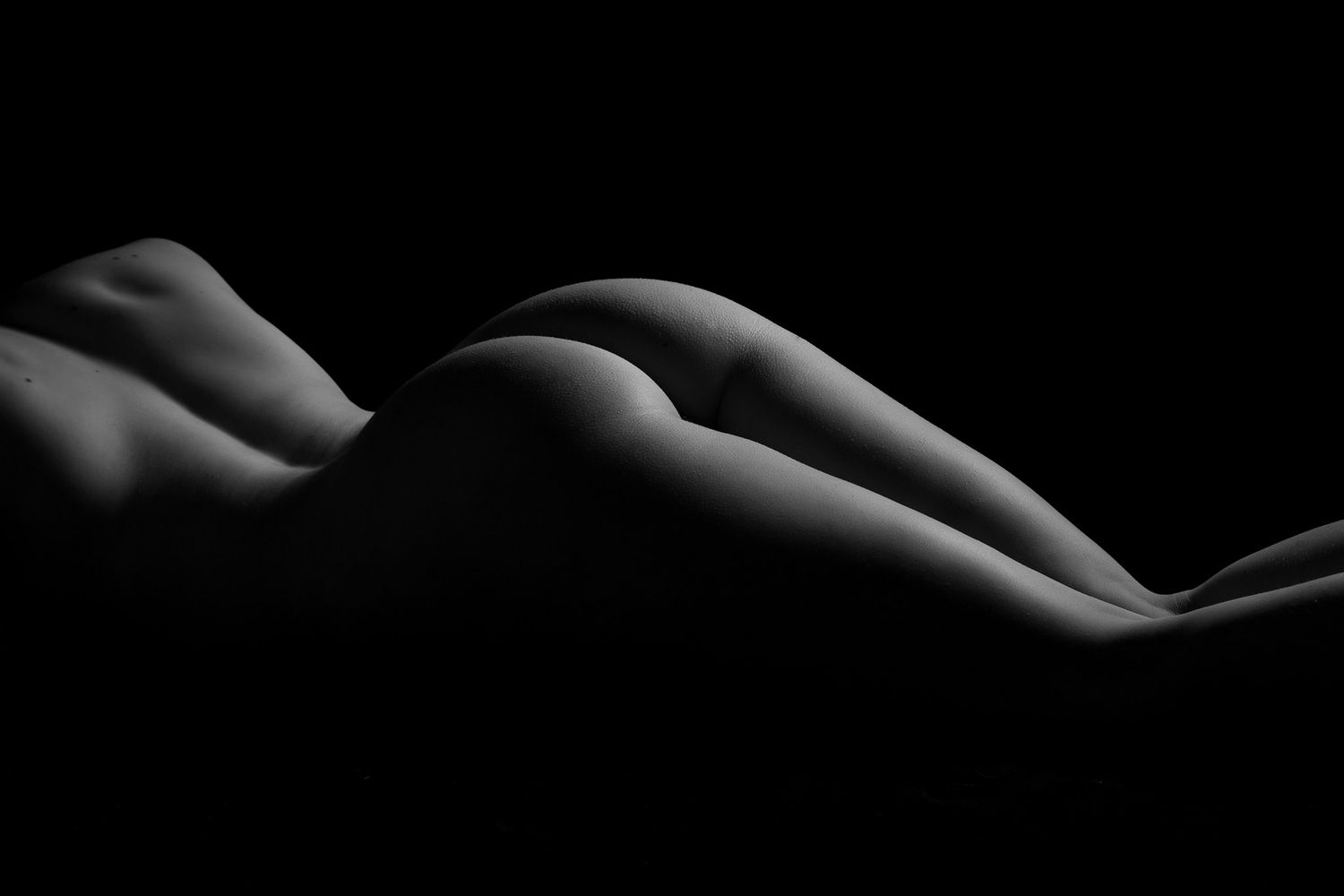 Black and white bodyscape photo highlighting the curves of a reclining nude figure against dark background. Boudoir photography Christchurch