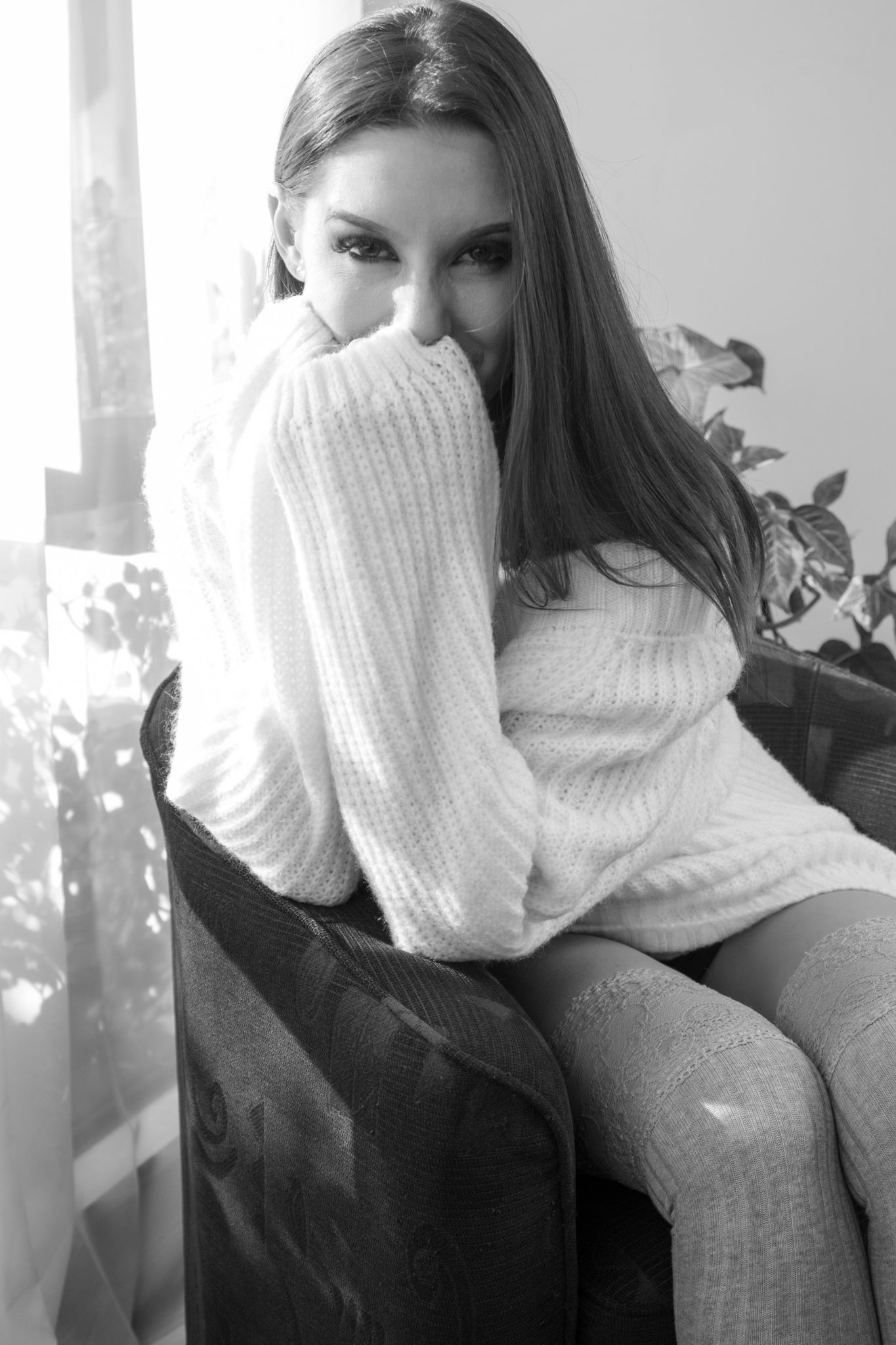 Young woman in cozy white knit sweater sitting in chair, smiling shyly while covering face near window. Boudoir photography Christchurch