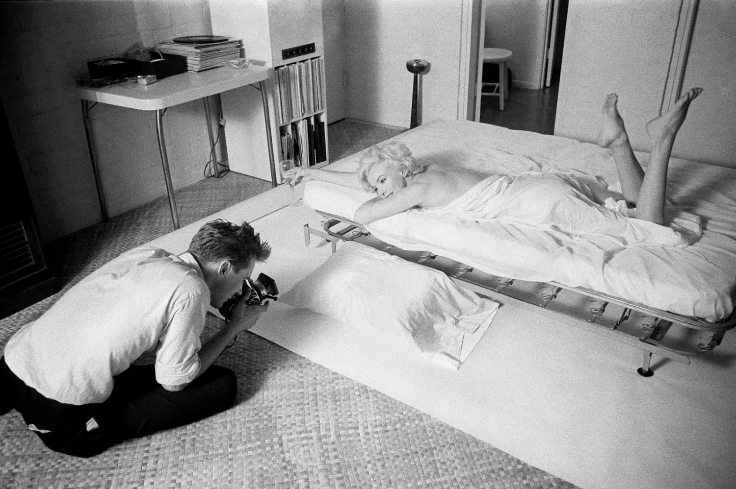 Black and white photo of a photographer kneeling to photograph a woman reclining on a bed in a bedroom.