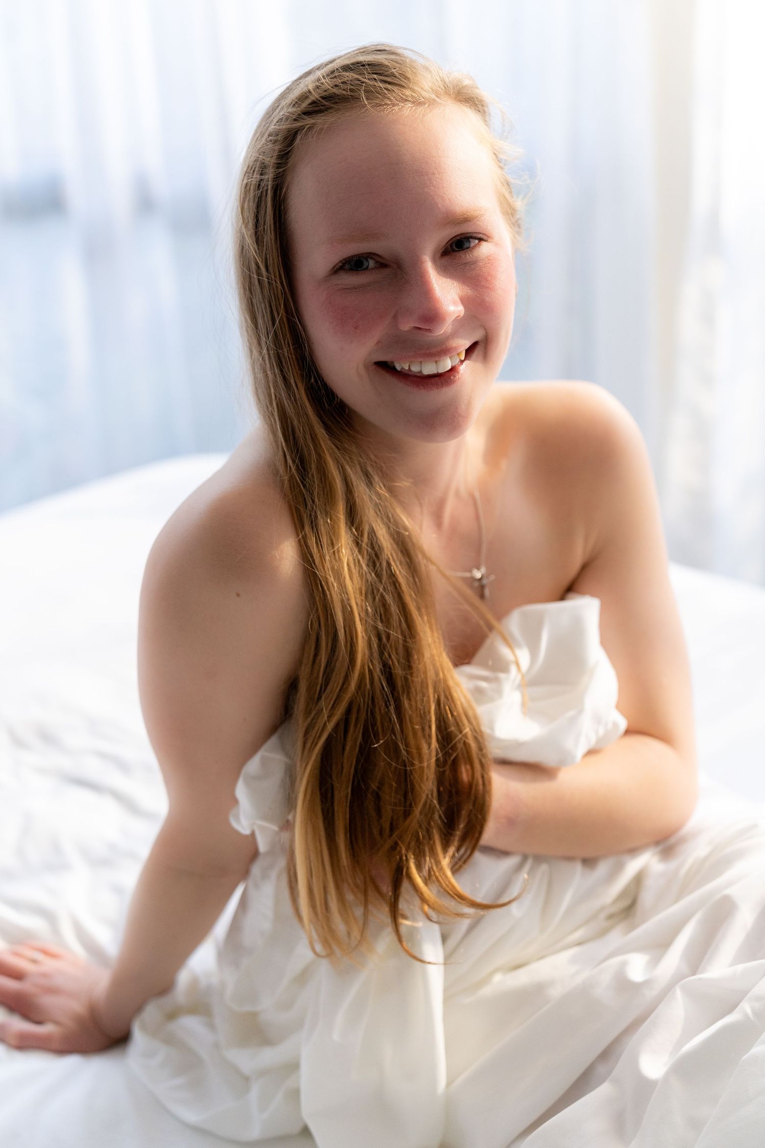 Smiling young woman with long hair wrapped in white sheets sitting on a bed near a bright window.