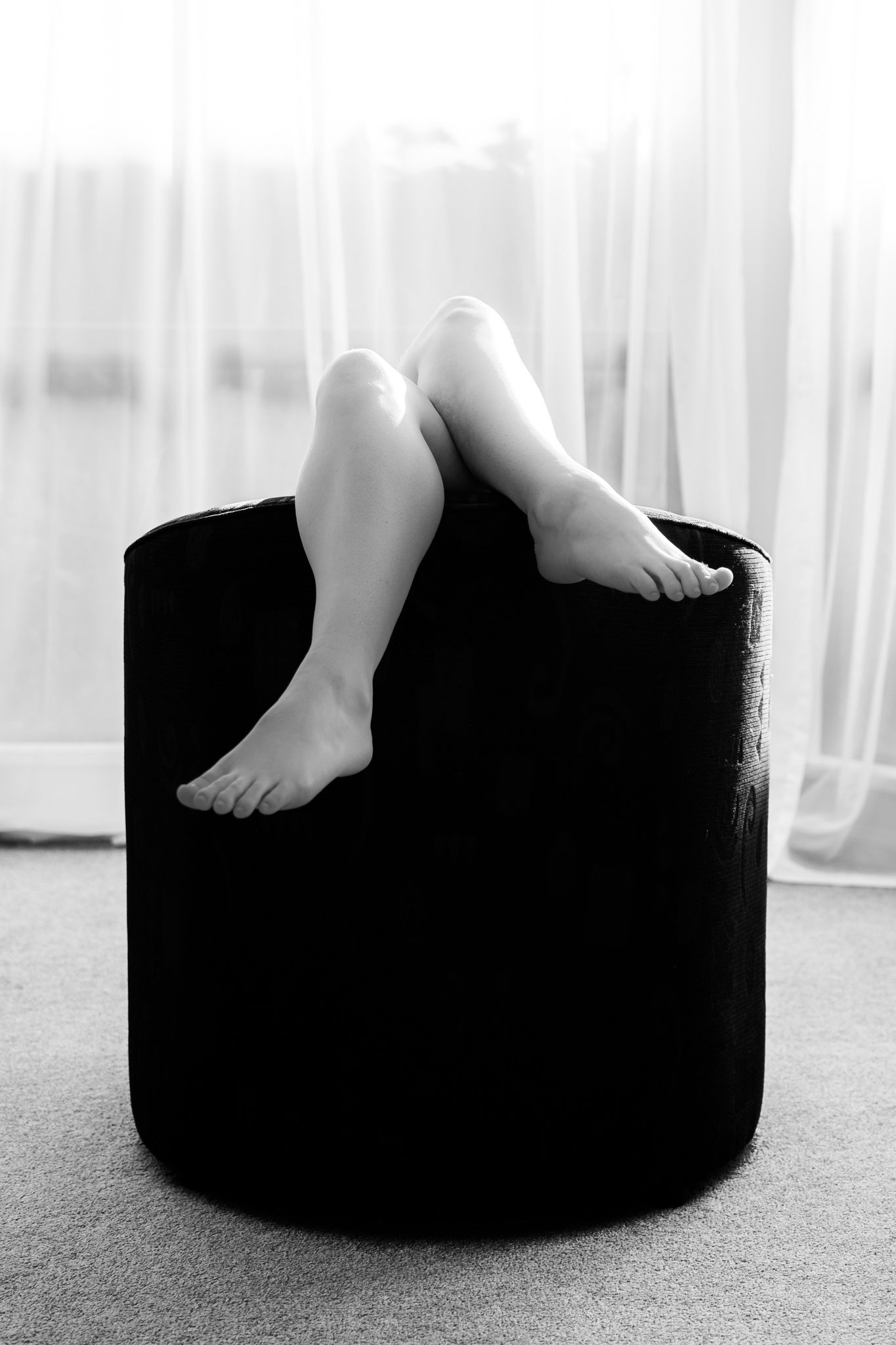 Black and white photo of bare legs and feet resting over the edge of a round black ottoman. Boudoir photography Christchurch