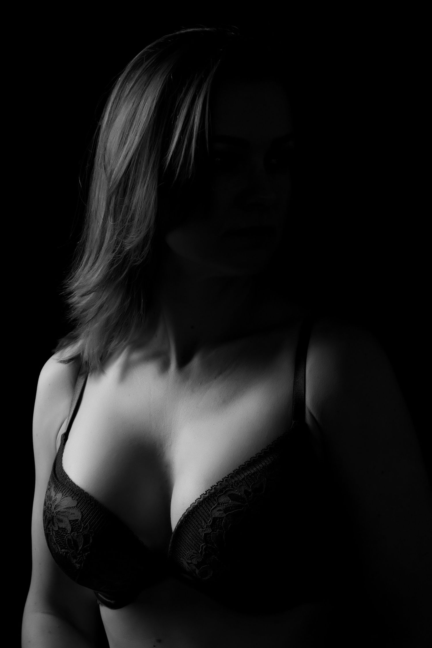 Dramatic black and white low-key portrait of a woman in a lace bra, partially shadowed against a dark background. Boudoir photography