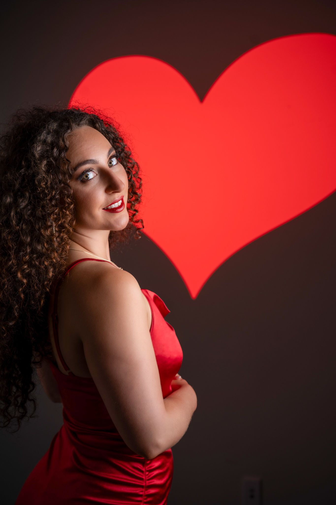 Woman in red satin dress poses confidently in front of a glowing red heart projection. Boudoir photography Christchurch portrait
