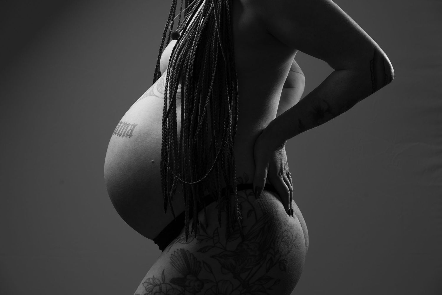 Black and white maternity photo of a pregnant woman with braids, showcasing her baby bump in a studio setting.