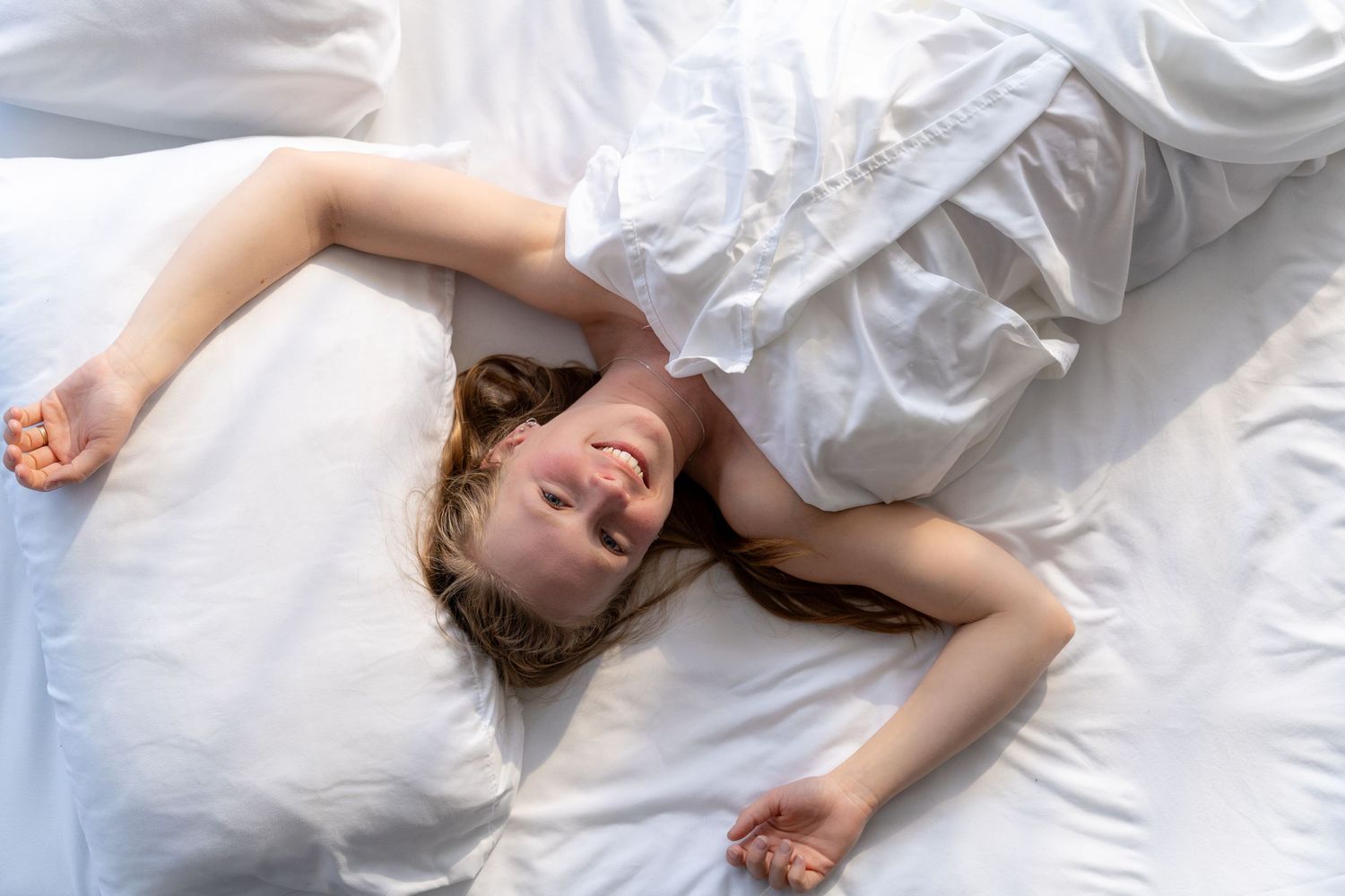 Happy young woman lying in white bed, stretching and smiling, enjoying a relaxing morning wake-up.