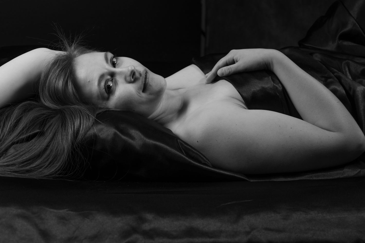 Black and white artistic boudoir photo of a woman lying on dark satin sheets with a contemplative gaze. Boudoir photography Christchurch