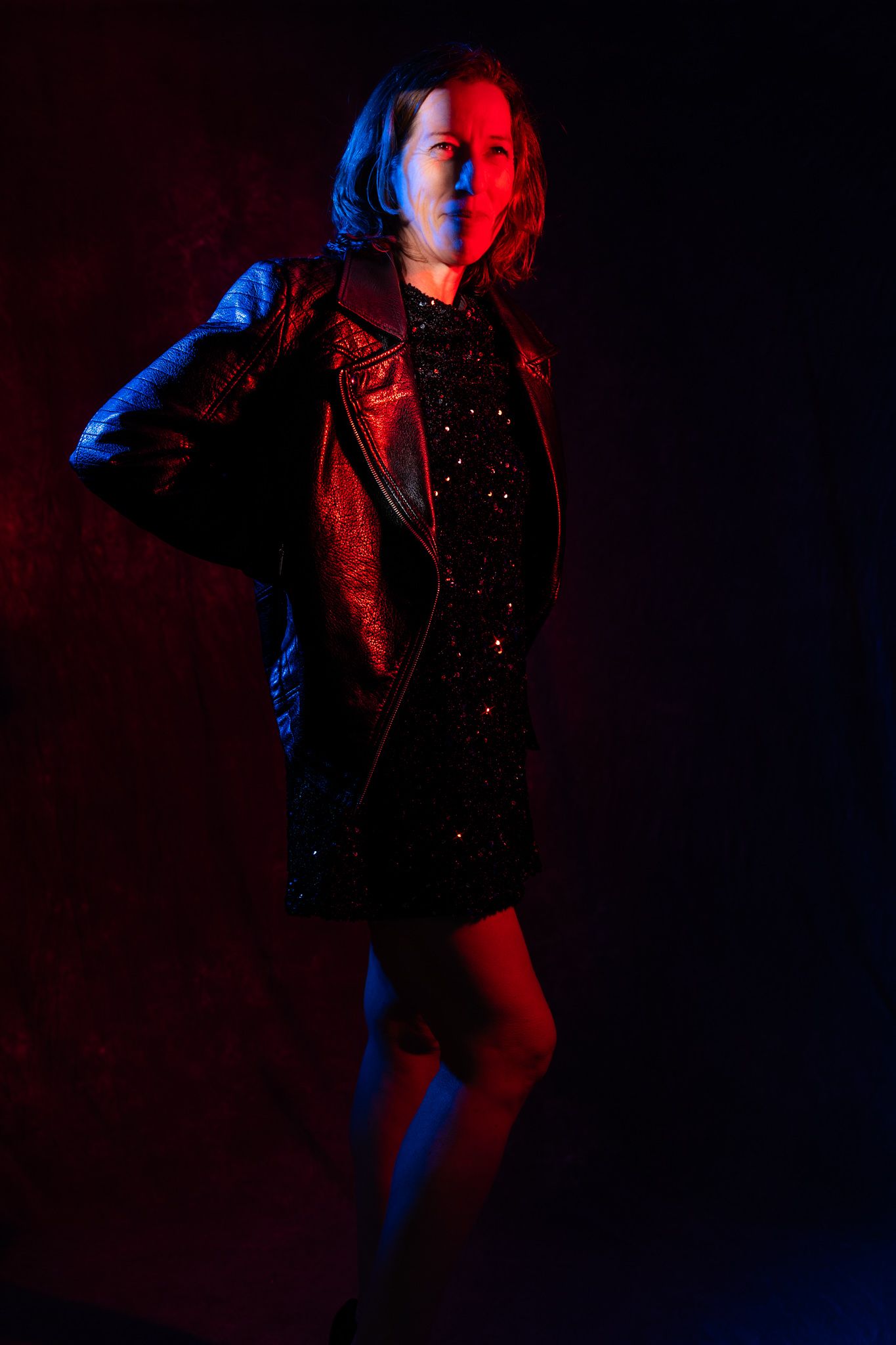 Woman in black leather jacket and sequin dress poses under dramatic red and blue studio lighting. Boudoir photography Christchurch portrait