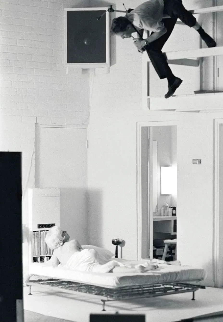 Black and white photo of a person leaping from a loft above a bed where another person lies sleeping.