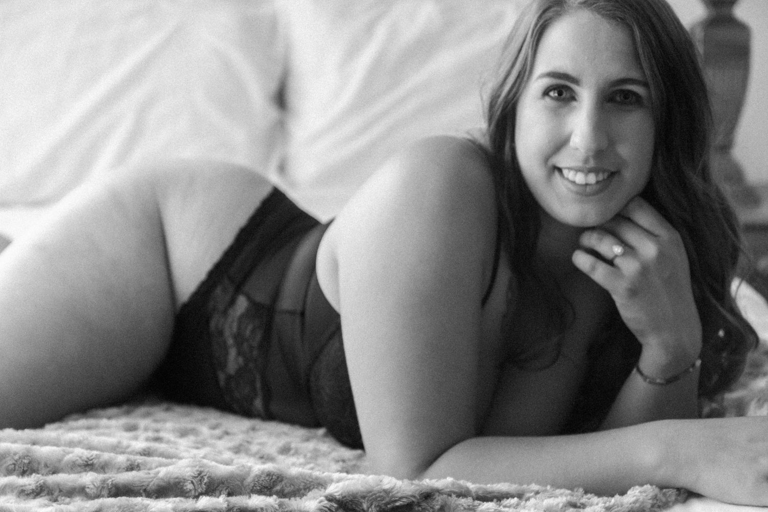 Black and white boudoir photo of a smiling woman in black lace lingerie lying on a bed. Boudoir photography Christchurch