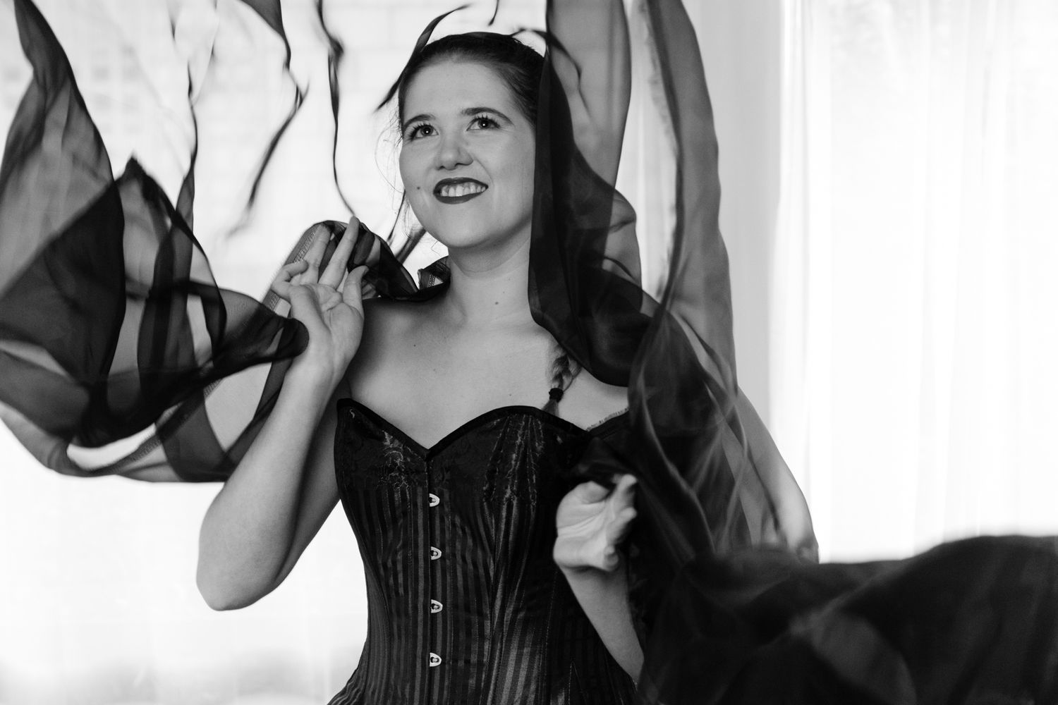 Black and white image of a smiling woman in a corset with flowing black fabric swirling around her. Boudoir photography Christchurch
