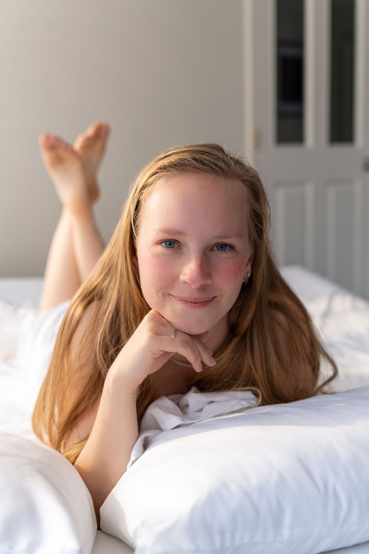 Young girl with long blonde hair lying on white bed, smiling at camera in bright bedroom.