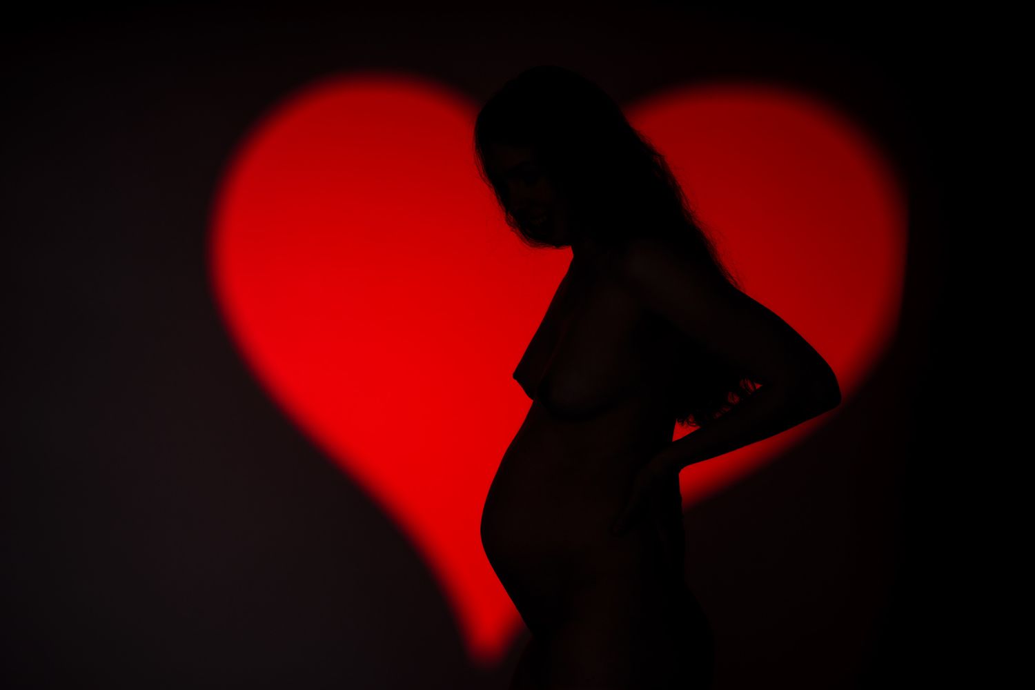 Silhouette of a pregnant woman against a red heart-shaped light projection on a dark background. Boudoir photography Christchurch maternity