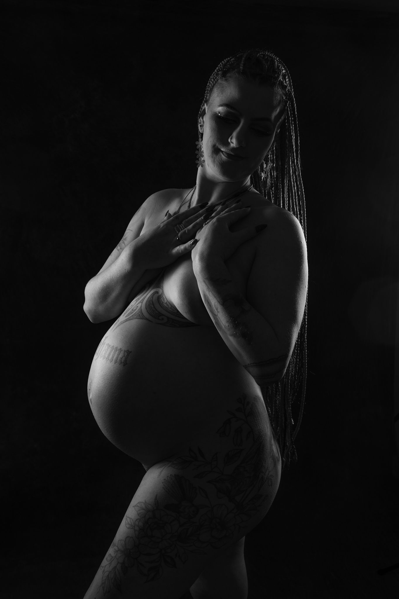 Black and white silhouette portrait of a pregnant woman with braids posing gracefully against dark background.