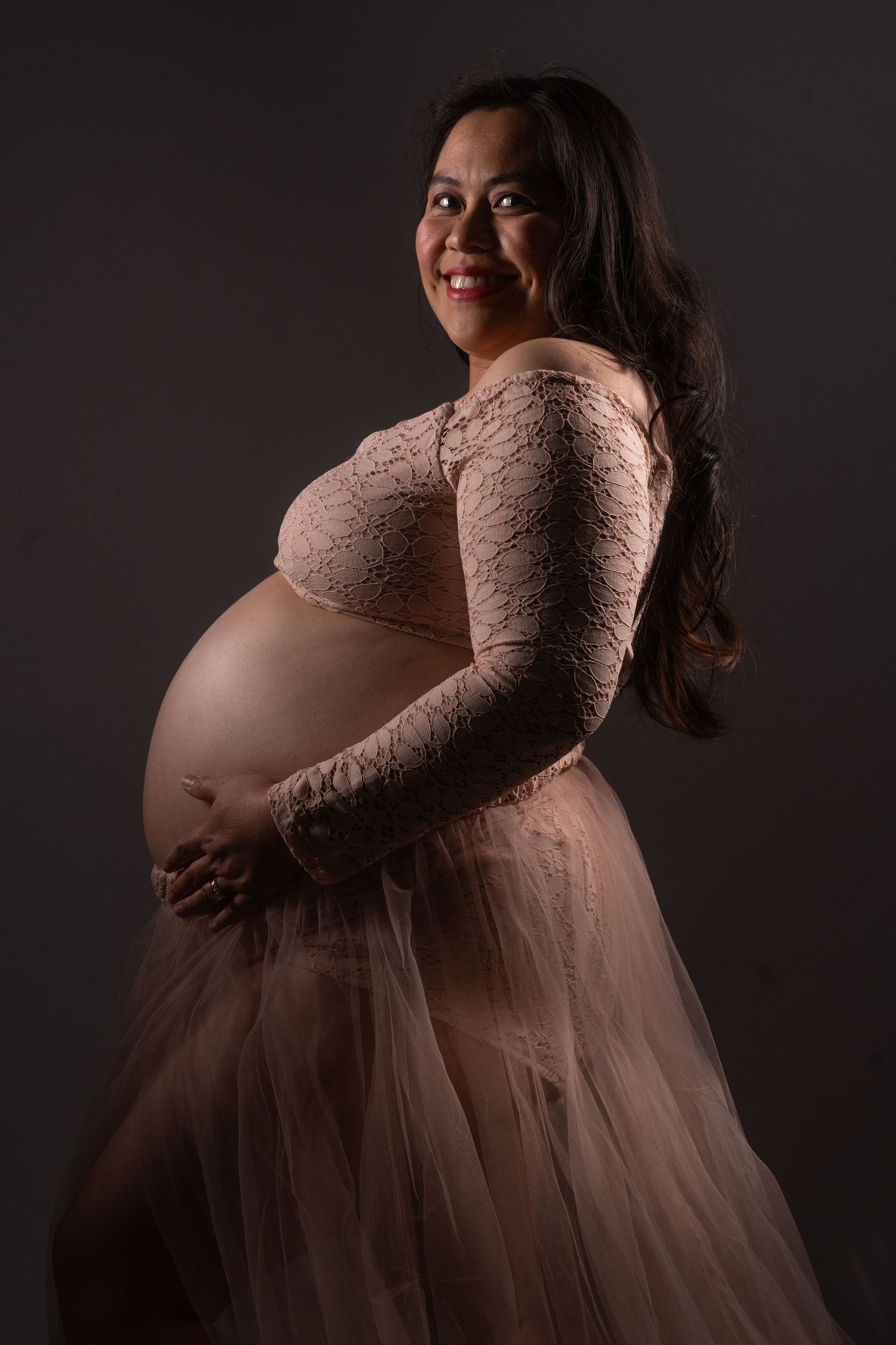 Smiling pregnant woman in pink lace maternity gown cradling her baby bump in studio. maternity photography Christchurch