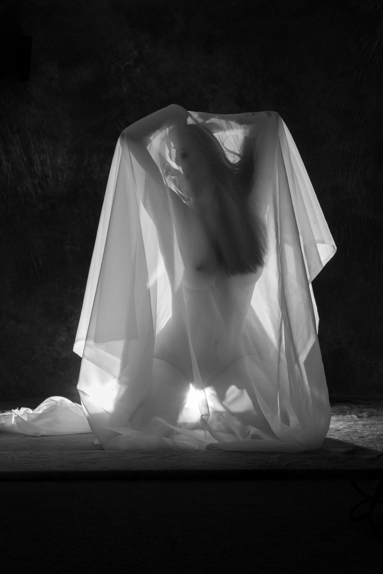 Artistic black and white nude silhouette of woman kneeling beneath translucent white fabric in dark studio. Boudoir photography Christchurch