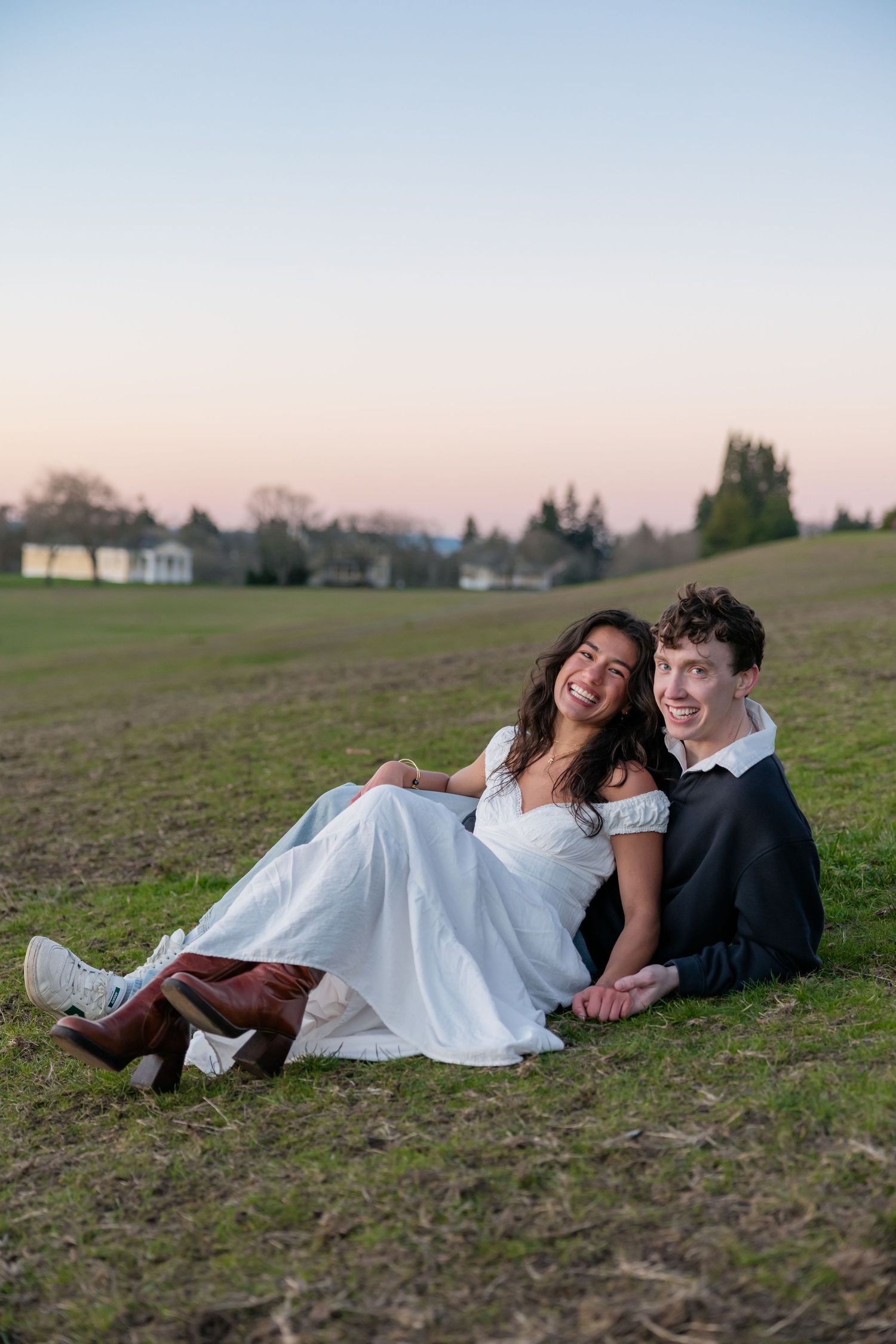 Happy couple sits together on grass at dusk, woman in white dress with brown boots, man in dark suit.
