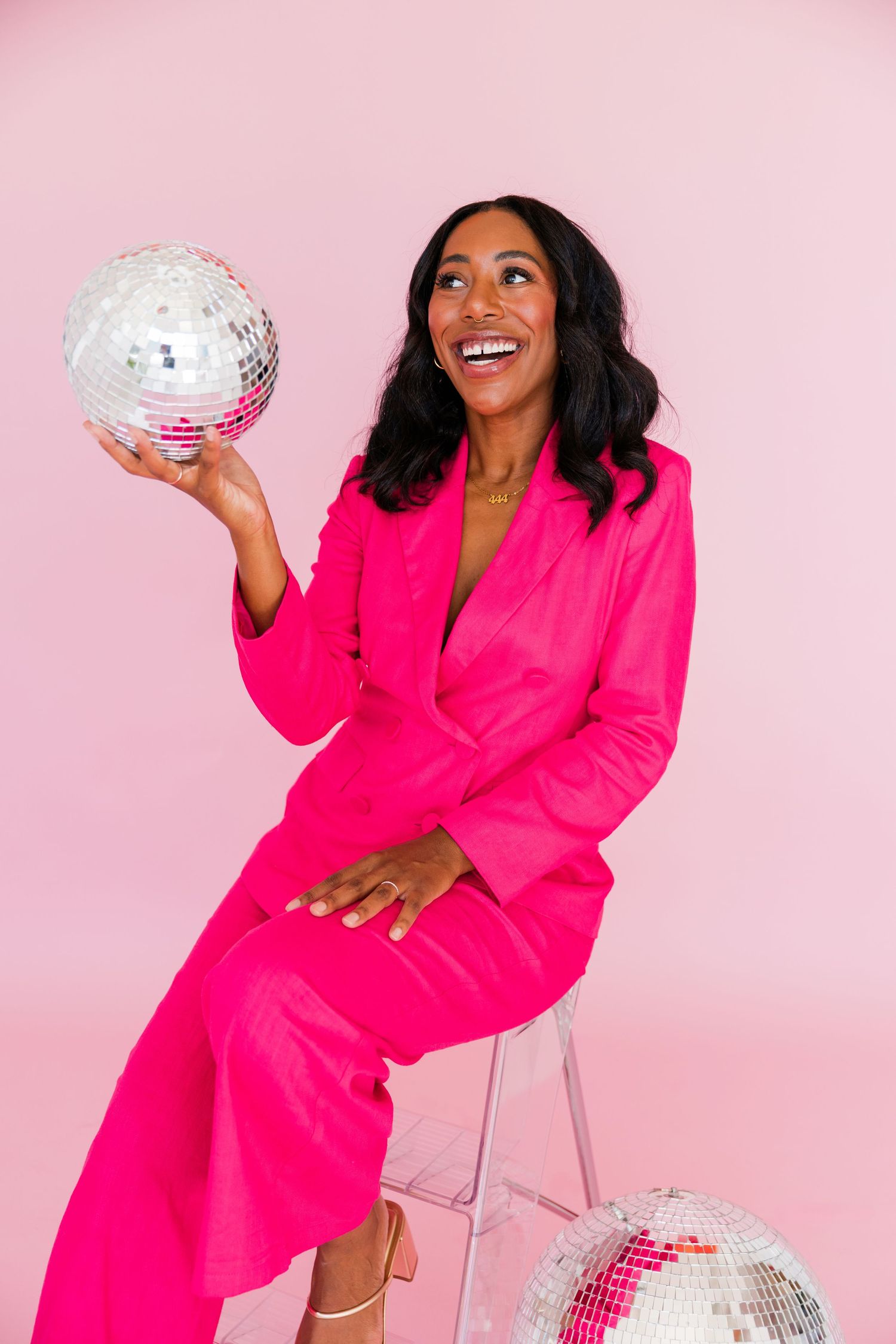 A joyful woman in a hot pink suit holds a disco ball against a pink background.