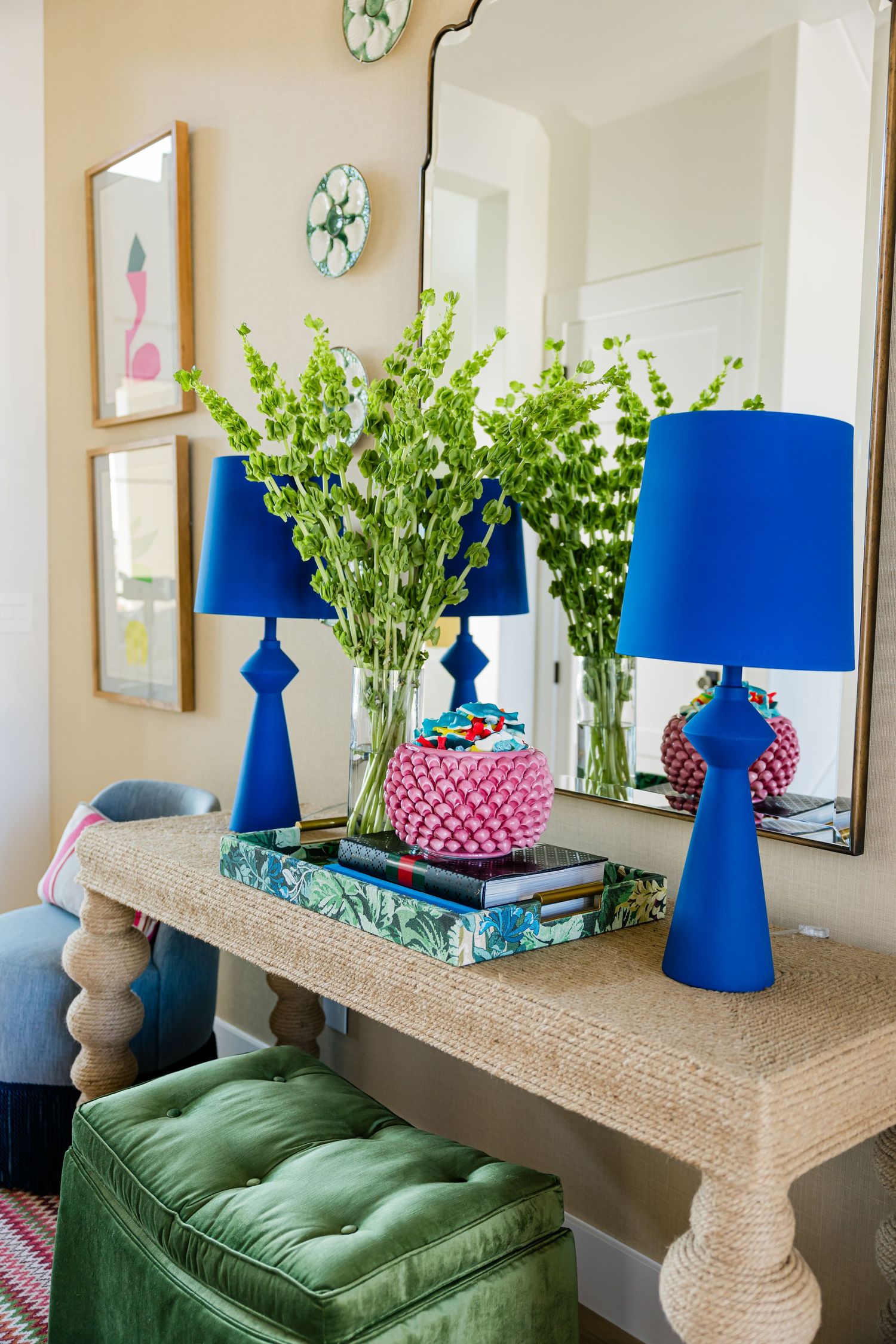 Rattan console table styled with cobalt blue lamps, green vases, and pink decorative object against beige wall.