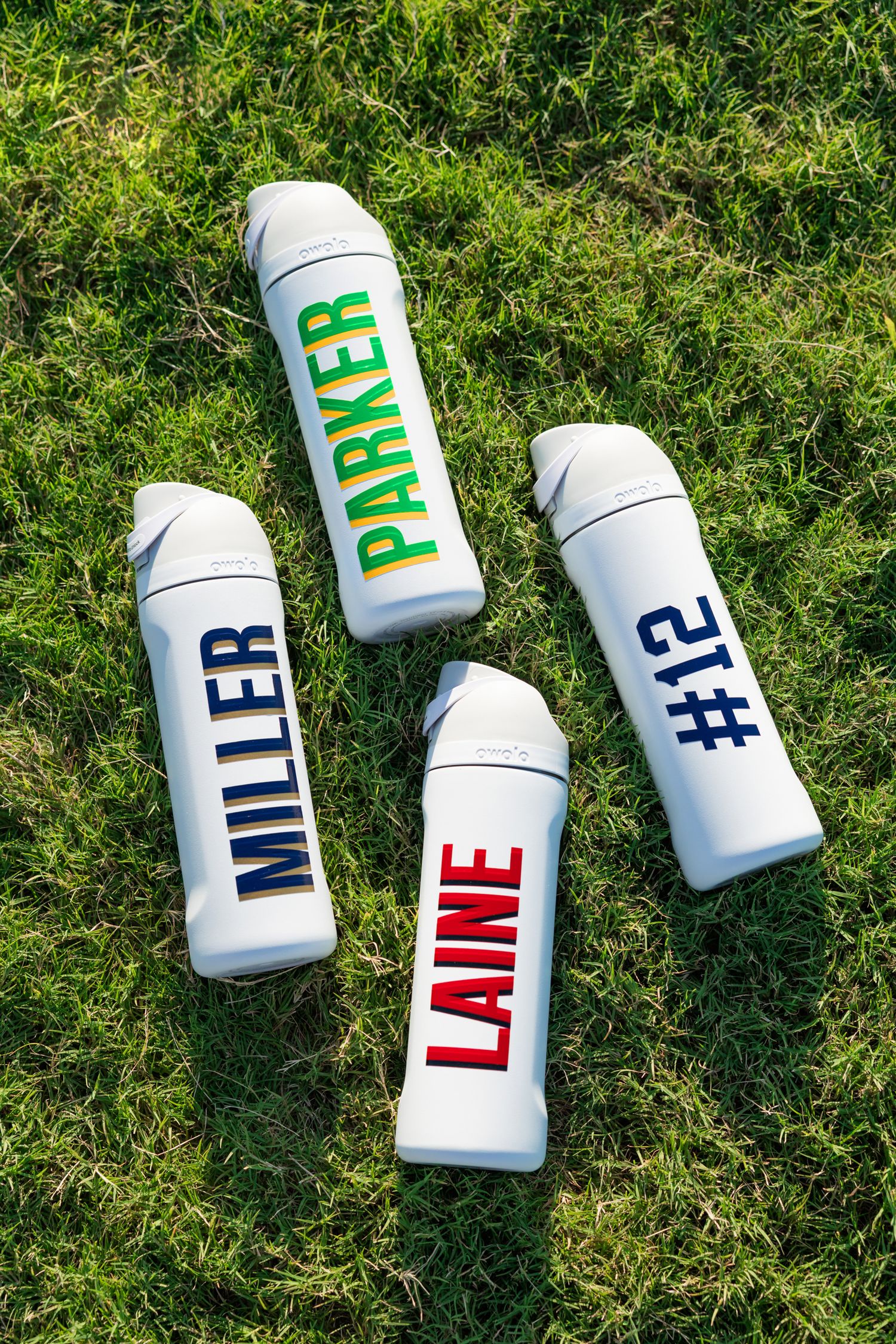 Four personalized white water bottles with names Miller, Parker, Laine, and number 12 on green grass.