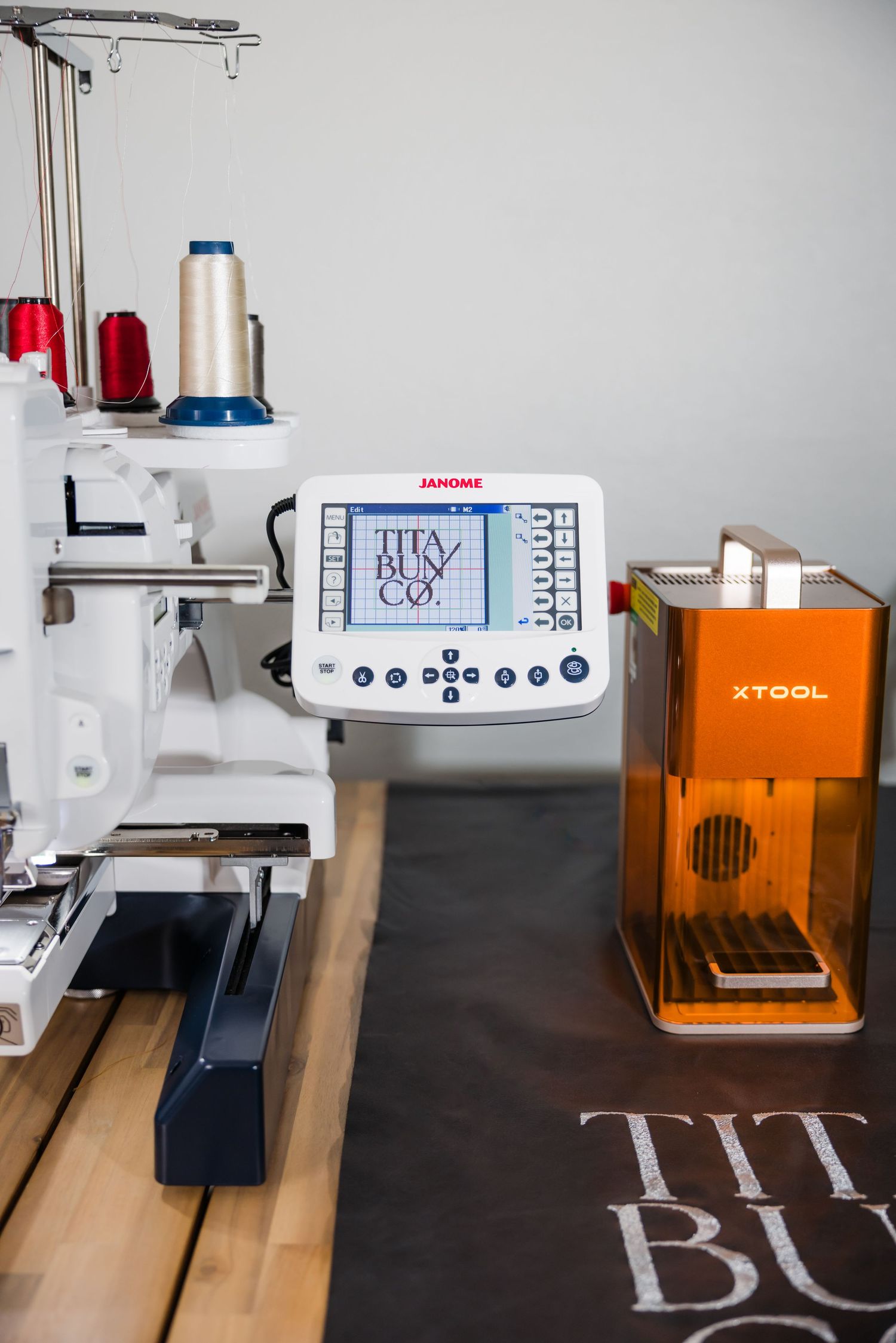 Janome embroidery machine with xTool laser engraver on wooden table, displaying Tita Bun Co. logo on screen.