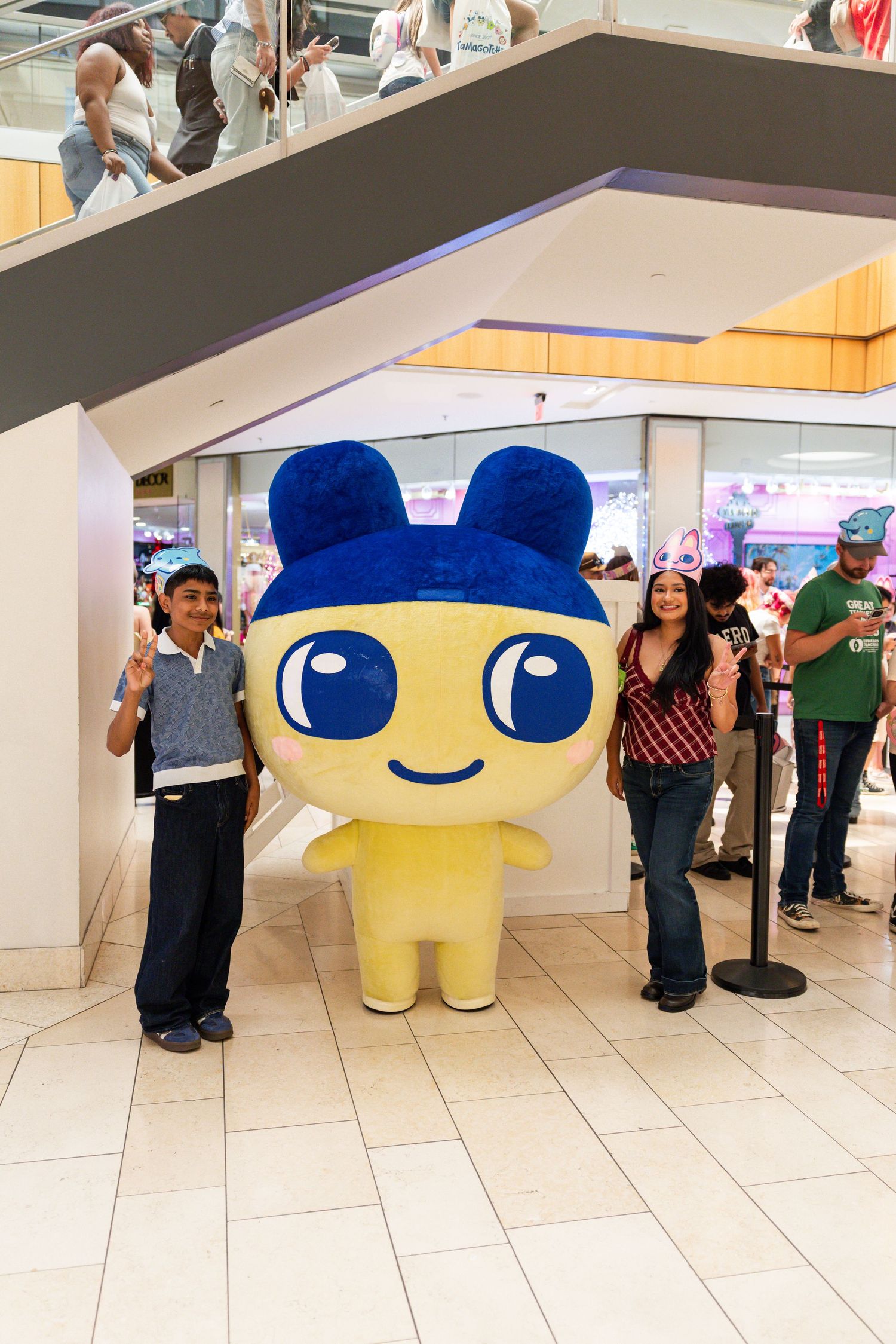 A boy and woman pose with a large Tamagotchi character mascot costume in a mall.