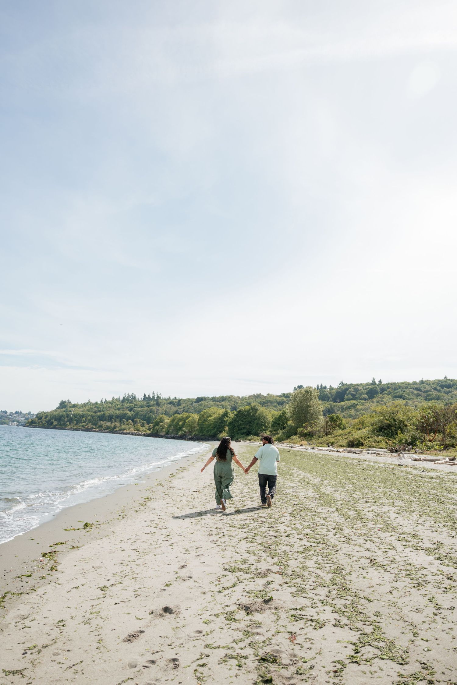 Couple holds hands walking away along a sandy beach shoreline with calm blue water beside them.