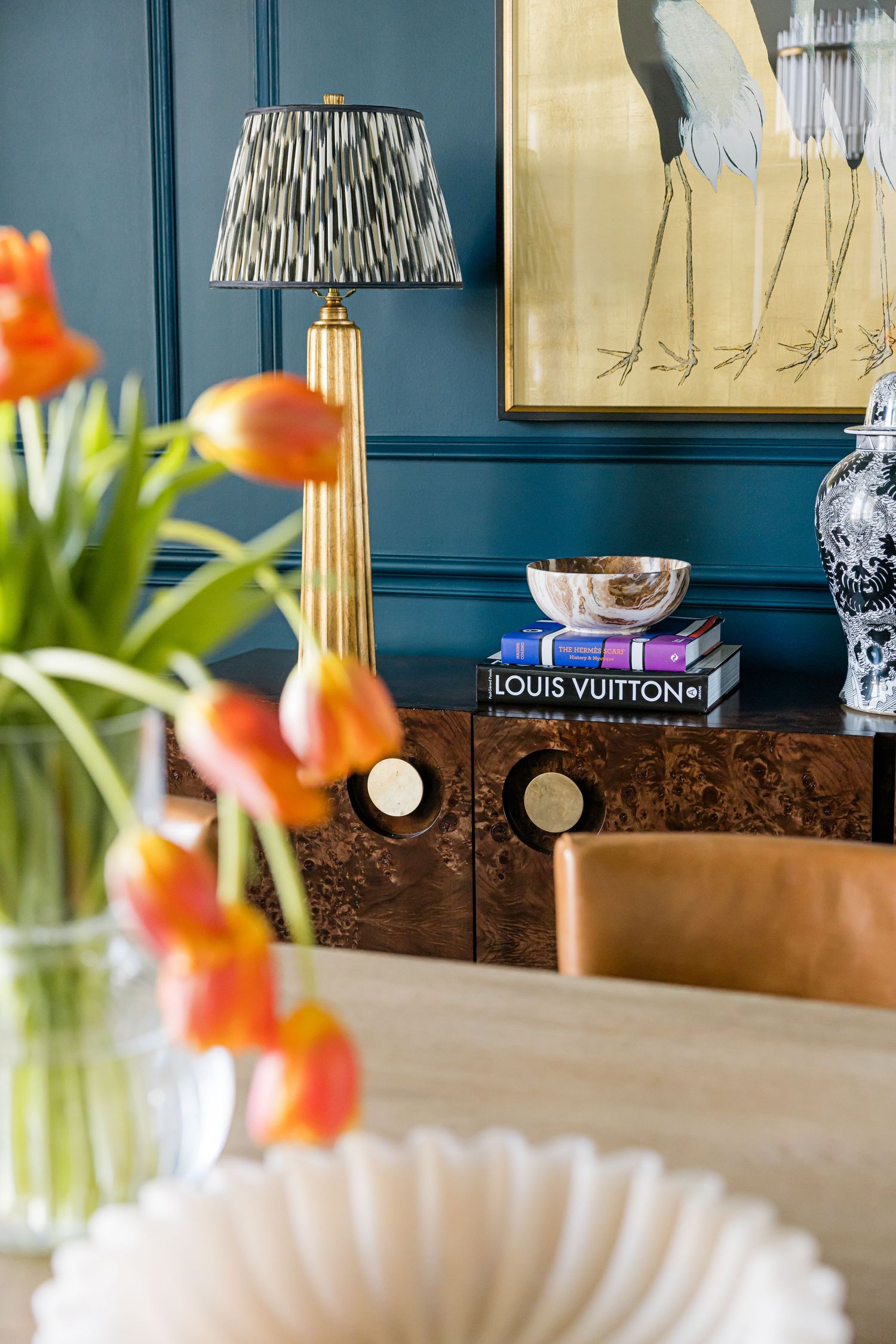 Close-up of a sideboard with Louis Vuitton books, silver bowl, gold lamp, and crane artwork on teal wall.