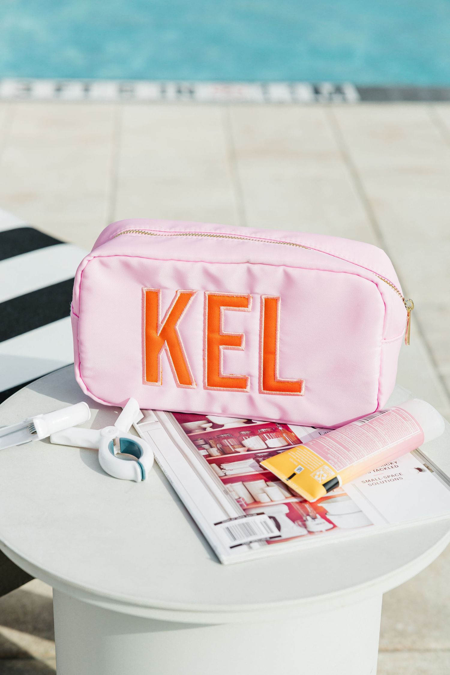 Pink monogrammed KEL nylon cosmetic bag beside a magazine and sunscreen near a swimming pool.
