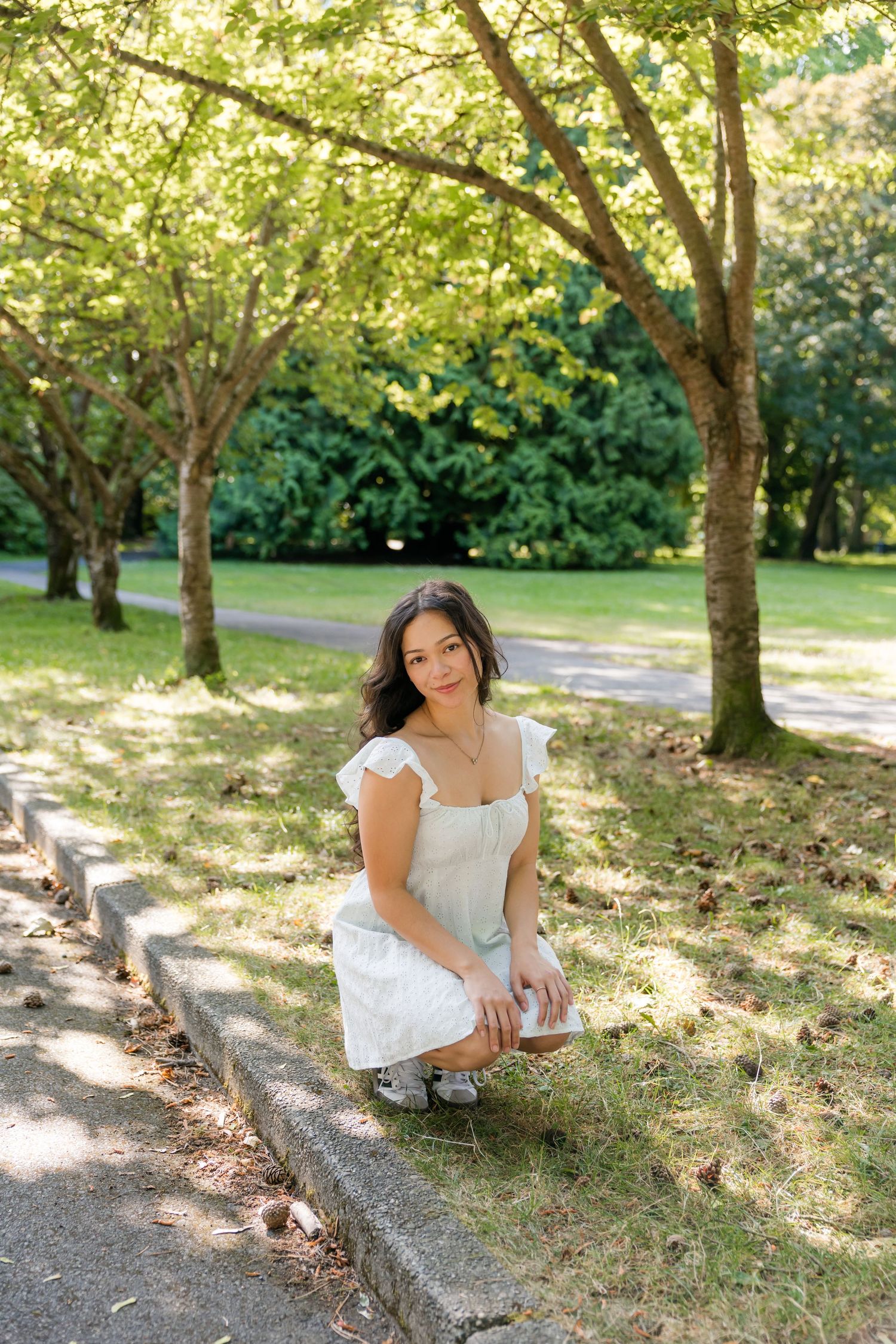 Young woman in white sundress crouches beside a park curb surrounded by lush green trees.
