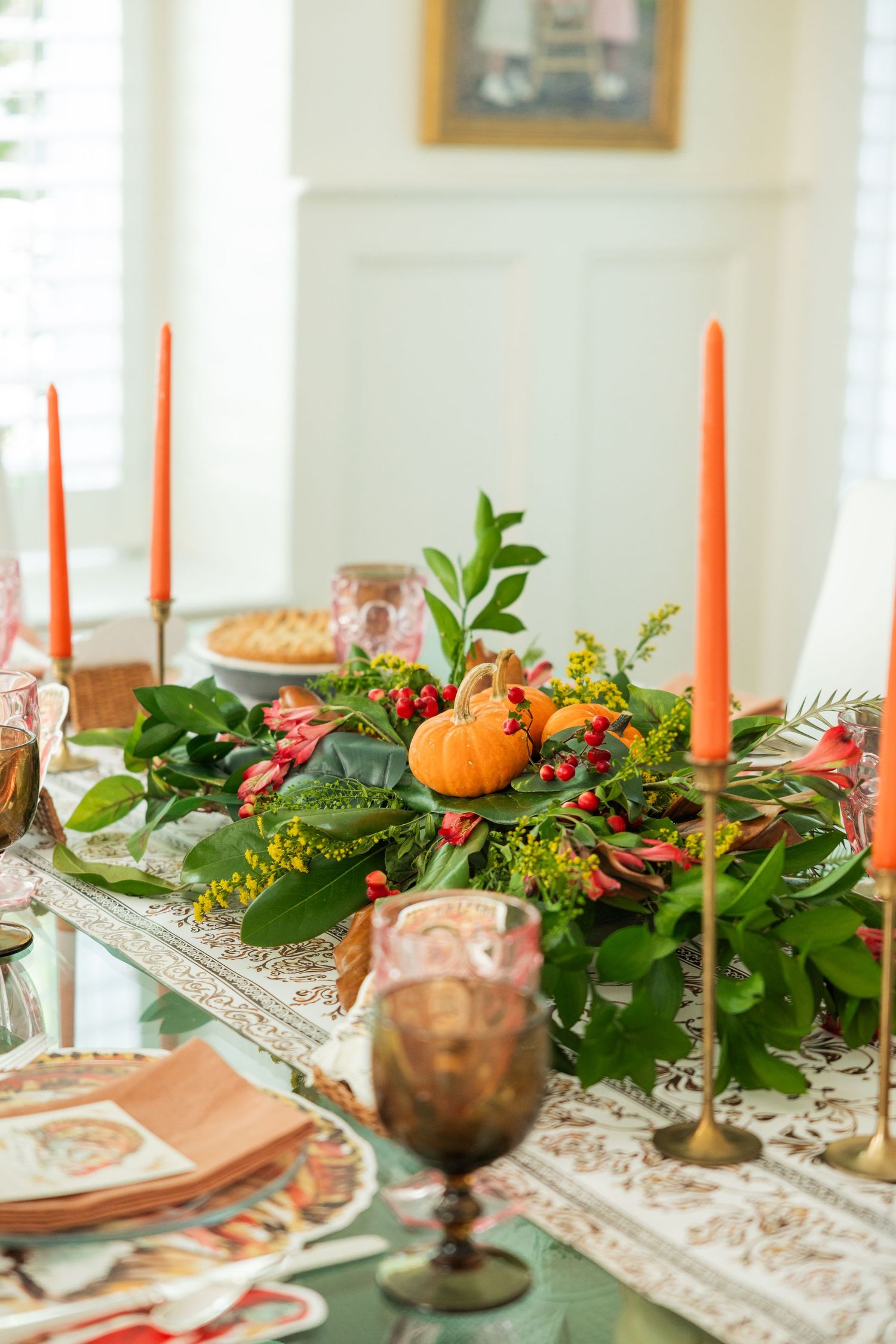 Elegant fall table centerpiece with greenery, mini pumpkins, berries, and tall orange taper candles.