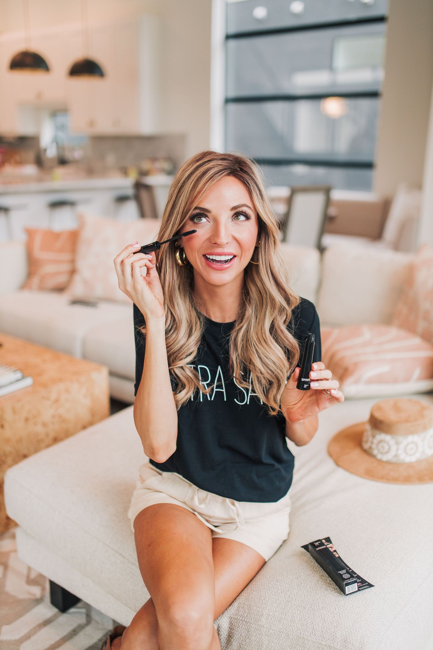 Smiling blonde woman applying IT Cosmetics mascara while sitting on white sofa in bright living room.