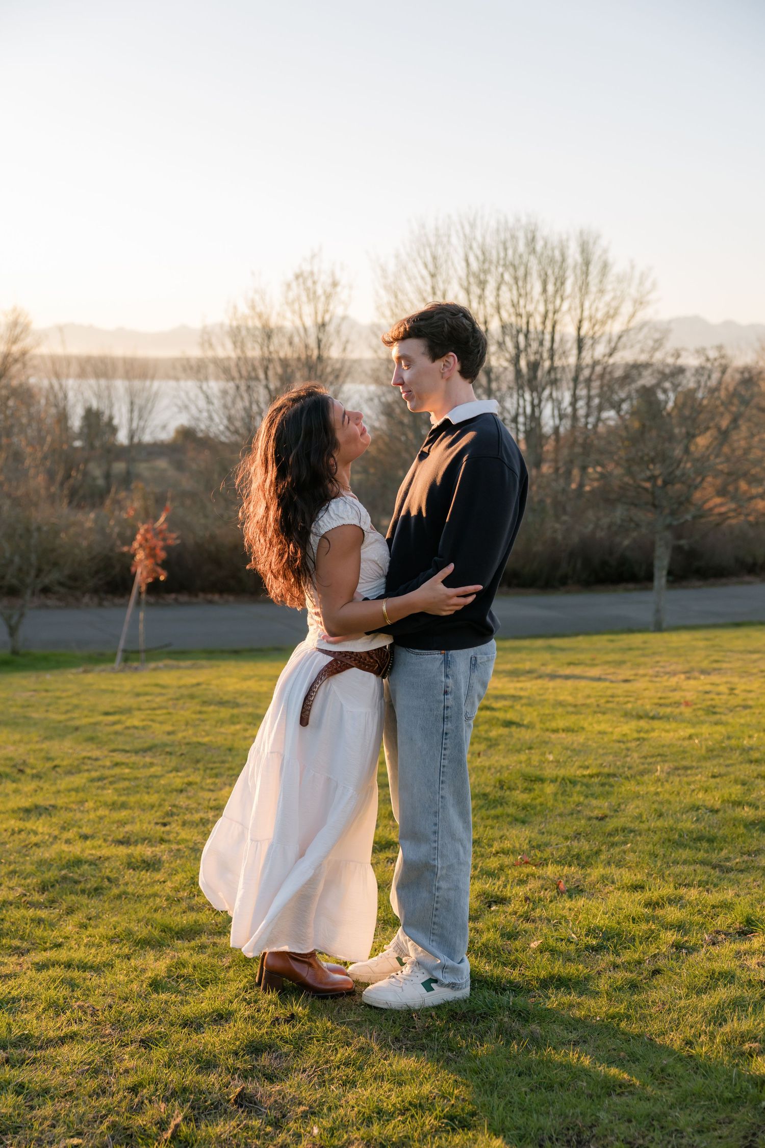A romantic couple embraces and gazes at each other in a sunlit park with mountains and water visible in the background.