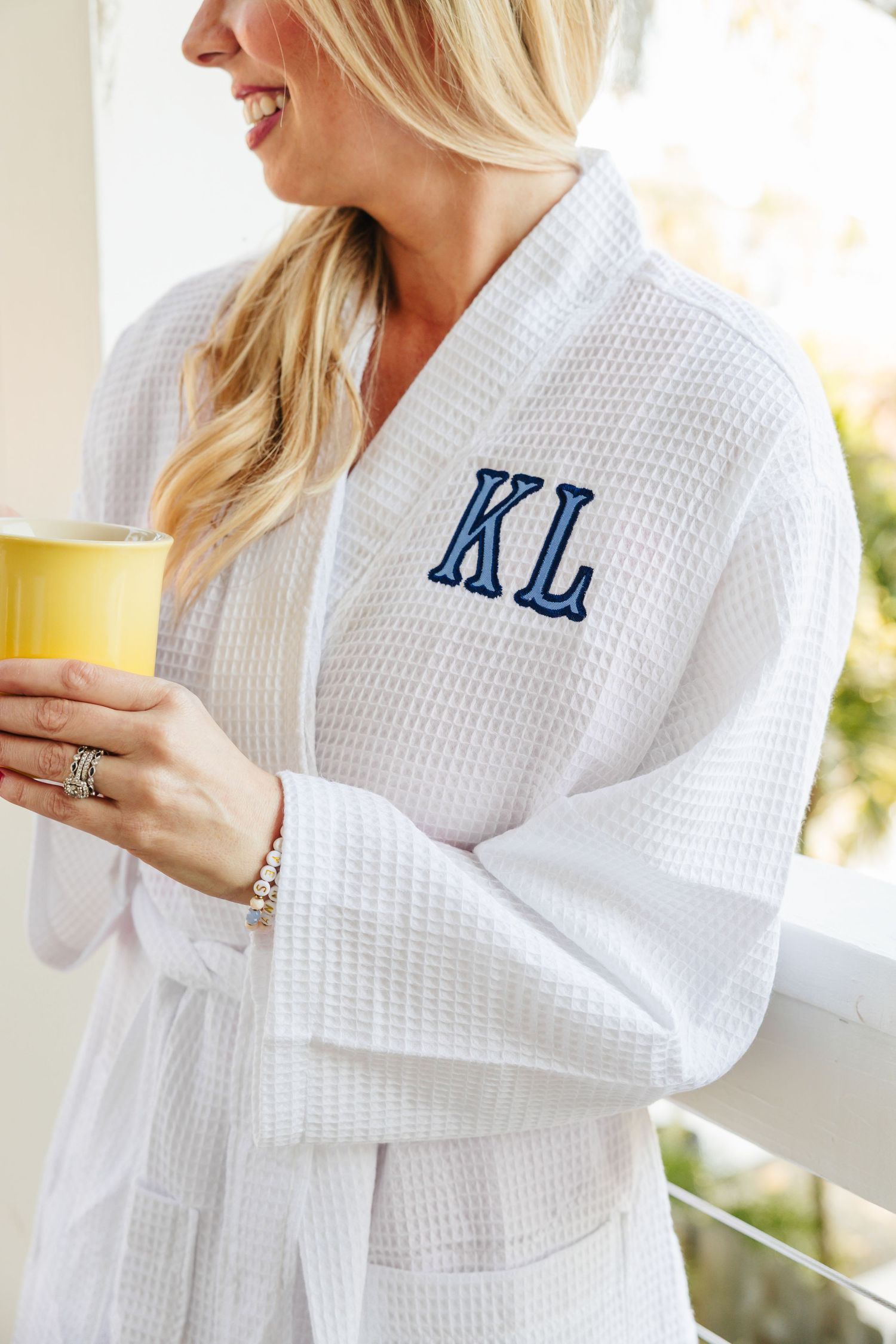 Blonde woman wearing a white waffle-knit robe with navy KL monogram holding a yellow mug.