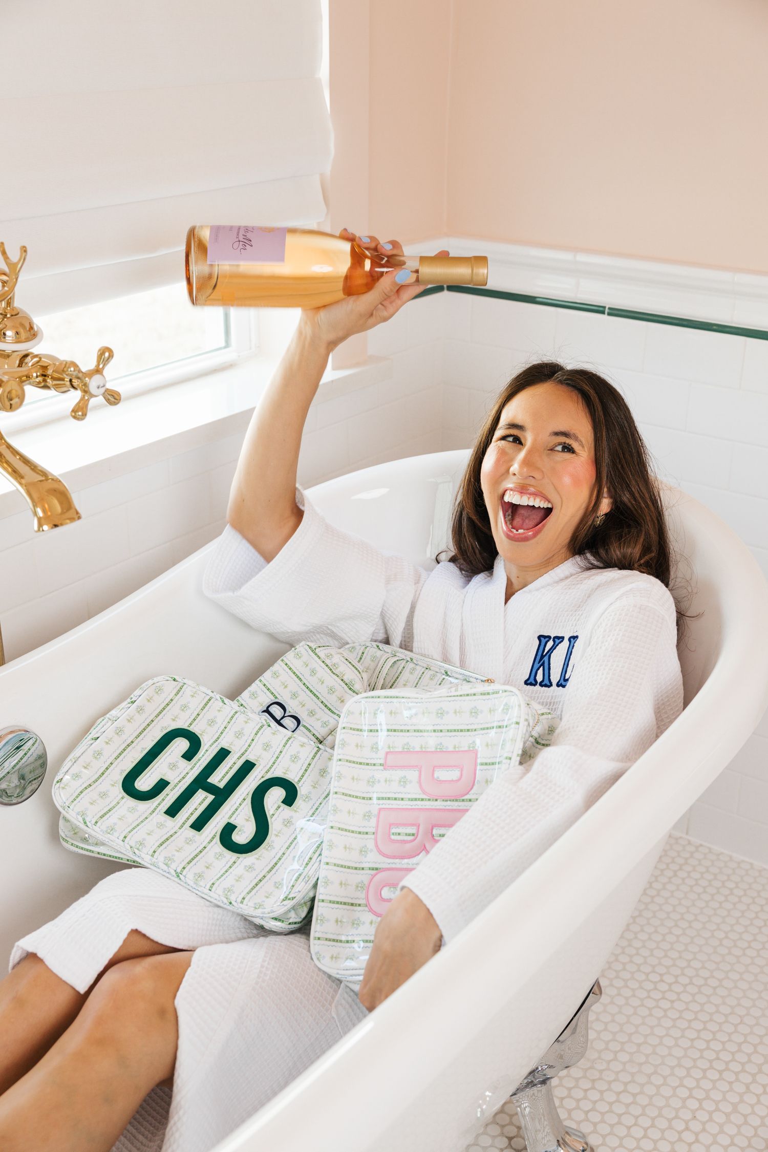 Woman in monogrammed robe holding rose wine bottle in clawfoot bathtub with CHS pillows.