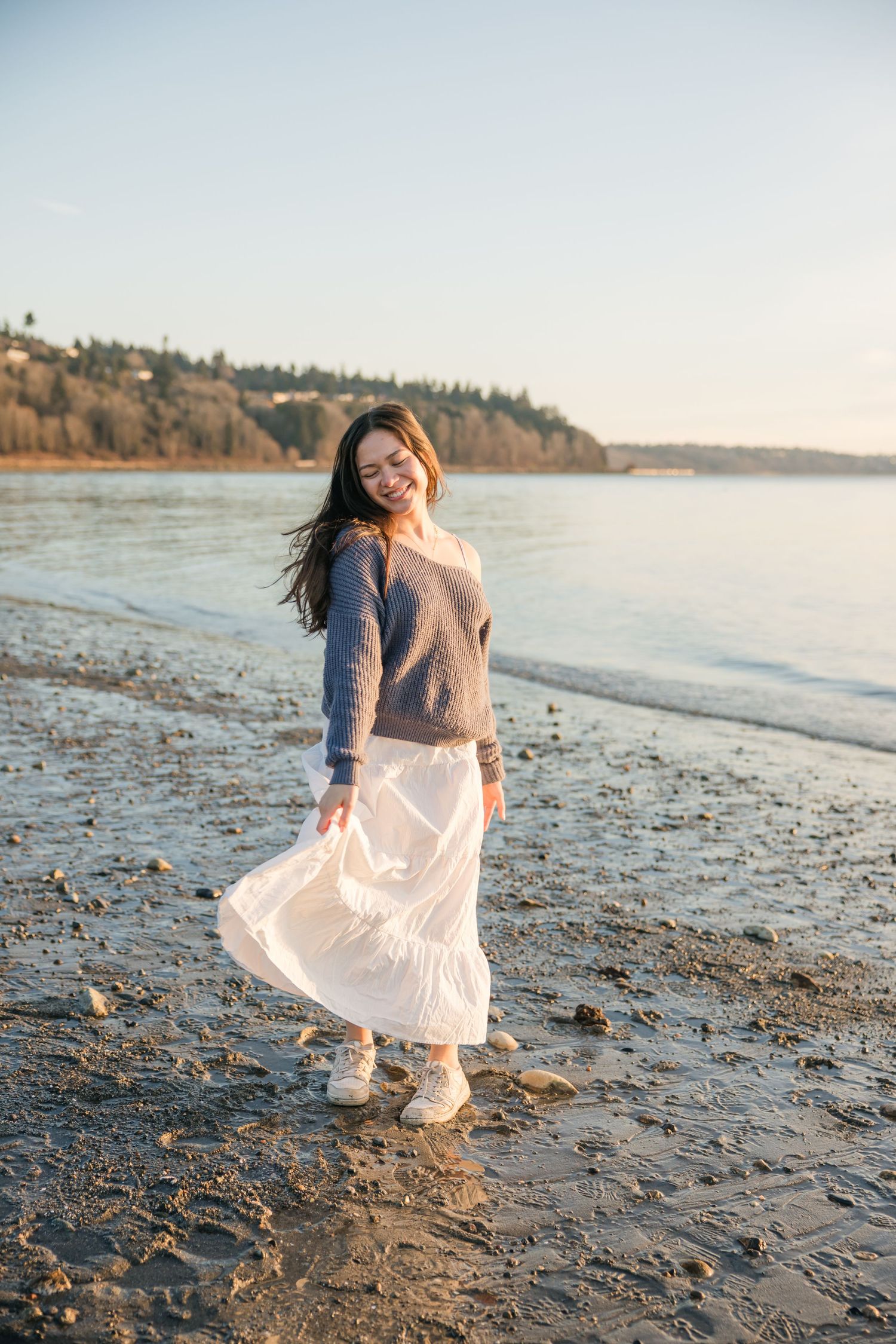 Smiling young woman in white skirt and gray sweater poses gracefully on a sunlit rocky beach.