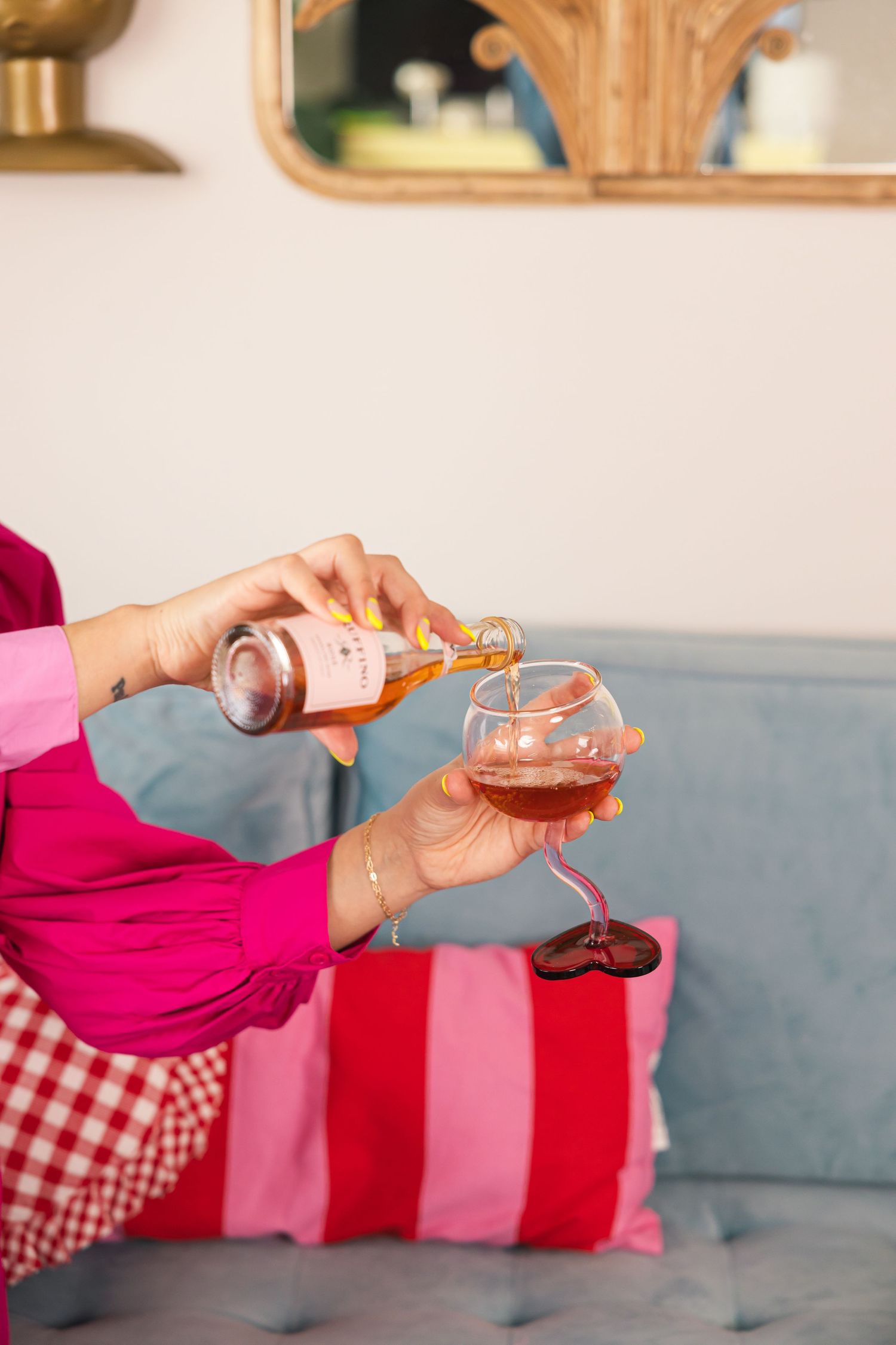 Woman in pink and magenta outfit pouring rose wine into a glass while seated on a blue sofa with colorful pillows.