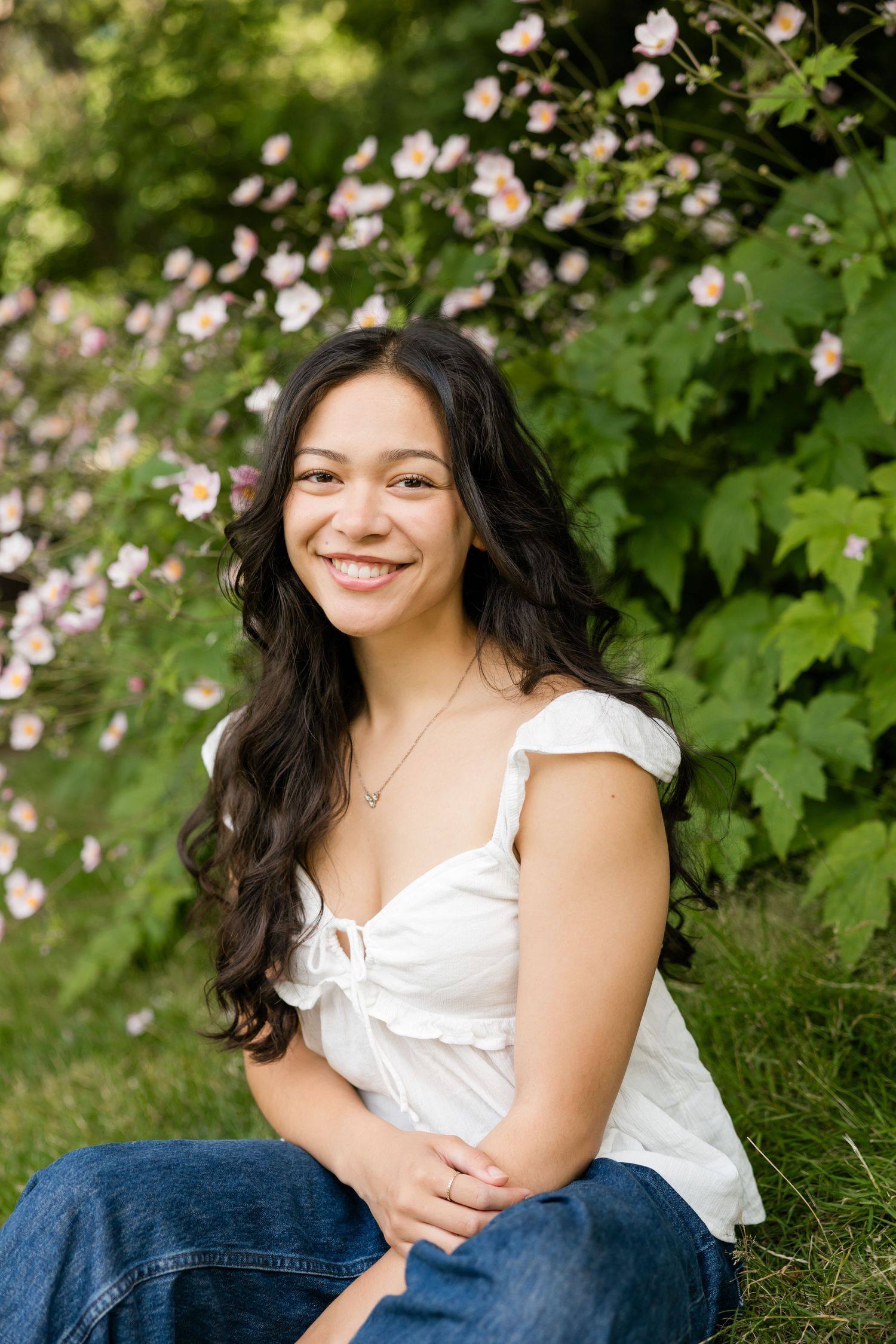 Smiling young woman with long dark hair sitting outdoors near blooming pink flowers in a white top and jeans.