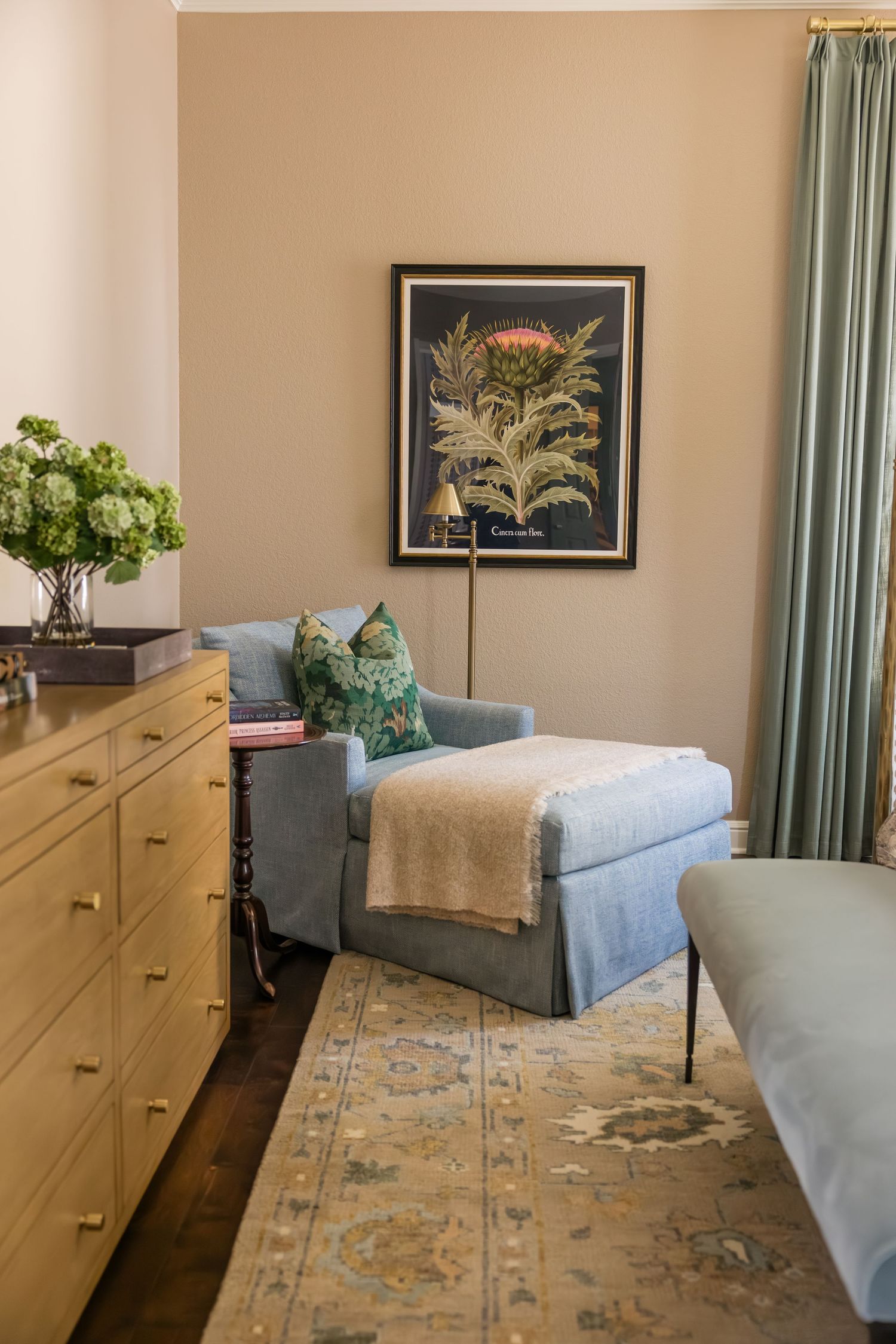 Cozy bedroom corner with blue chaise lounge, botanical art, light wood dresser, and floral area rug.