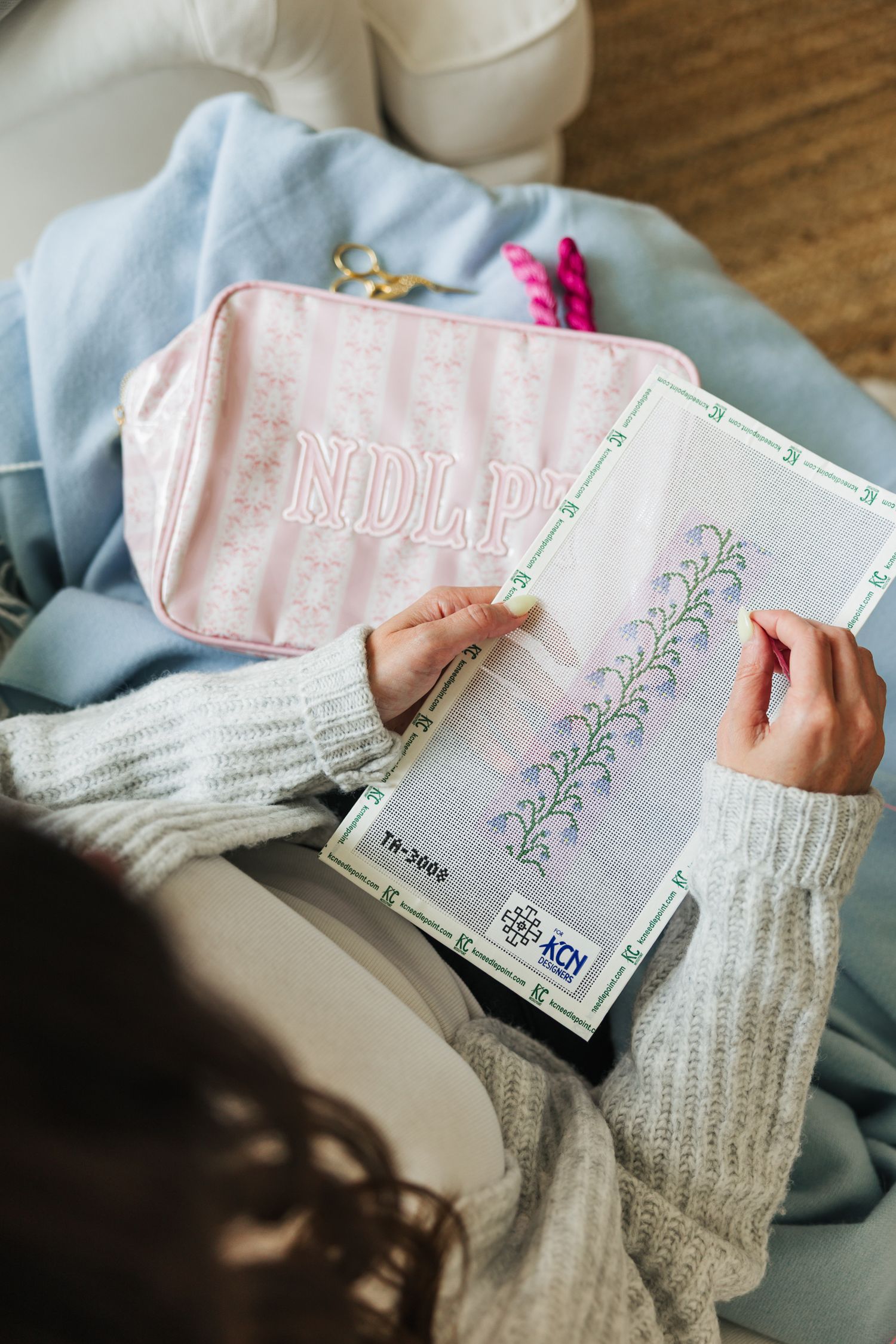 Woman doing needlepoint with a pink monogrammed pouch nearby on a cozy chair.