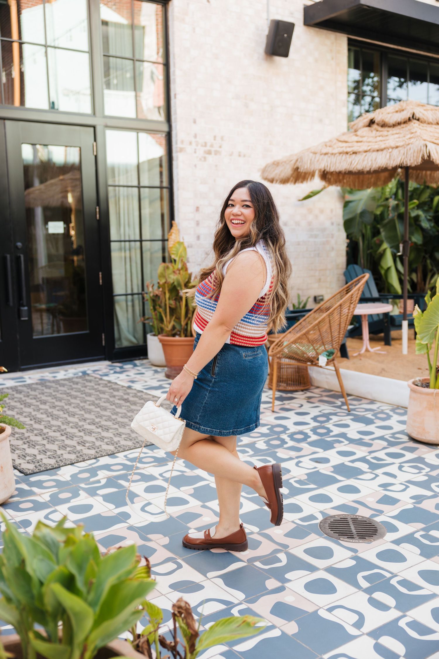 Smiling curvy woman in striped top and denim mini skirt with white bag and brown loafers on a patterned patio.