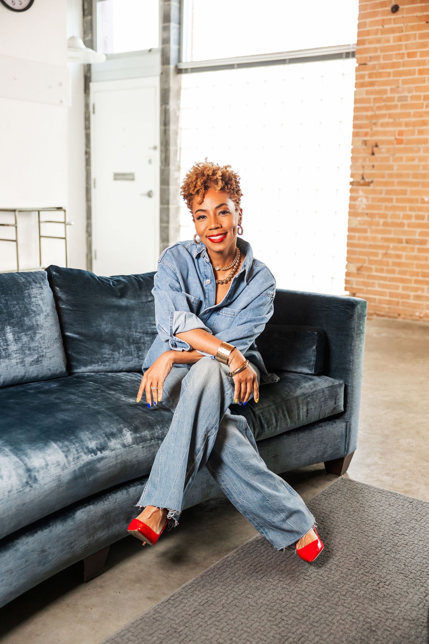 Stylish woman in denim outfit with red heels sitting on blue velvet sofa in loft.