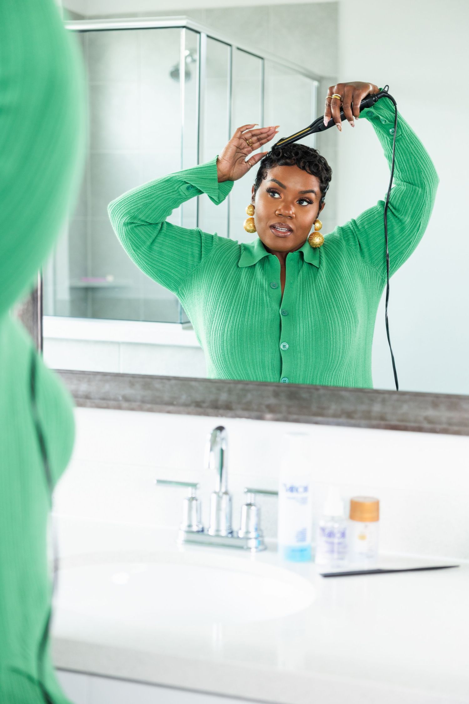 A woman in a green button-up shirt styling her short hair with a flat iron while looking in a bathroom mirror.