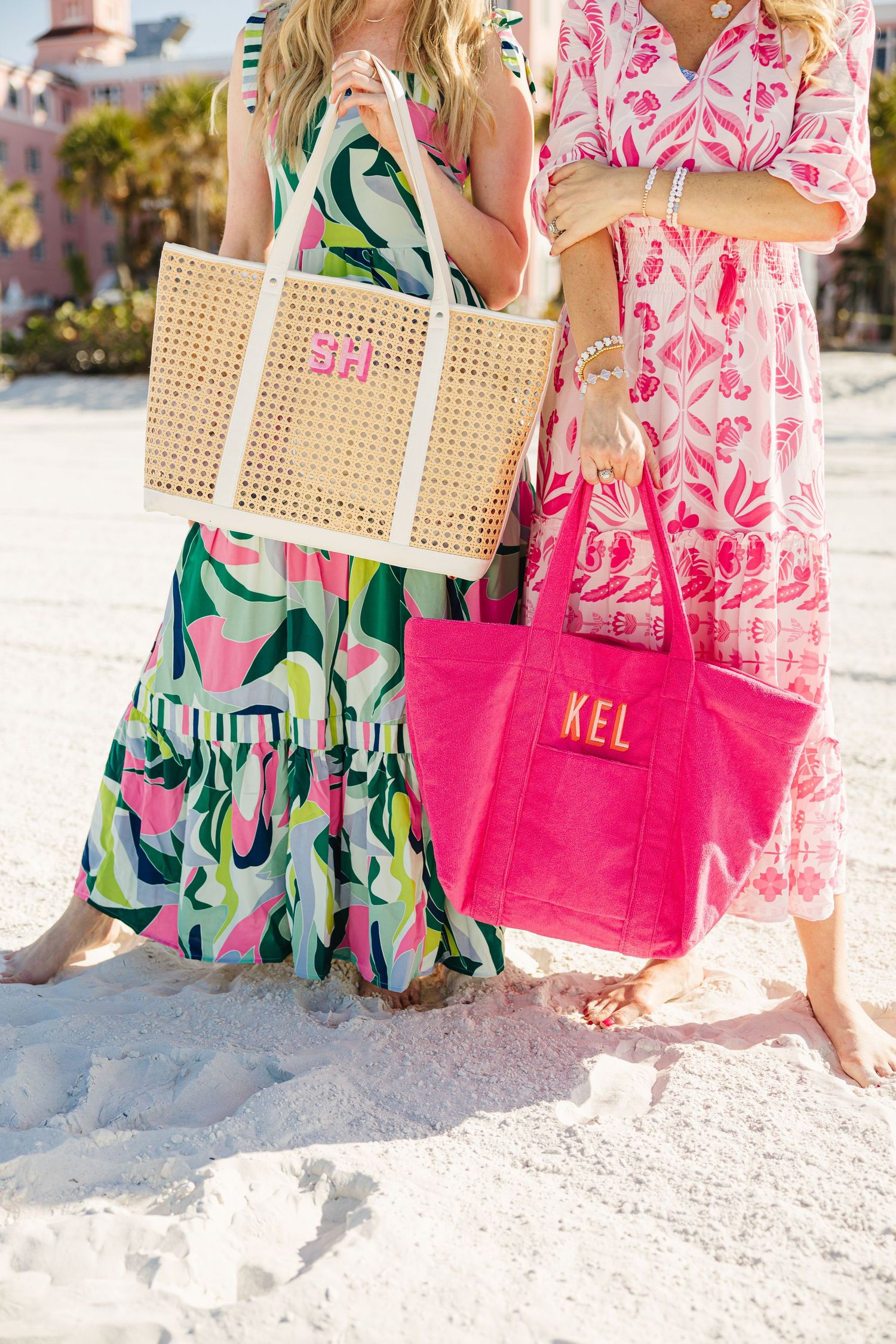 Two women holding monogrammed rattan and hot pink canvas tote bags on white sandy beach.