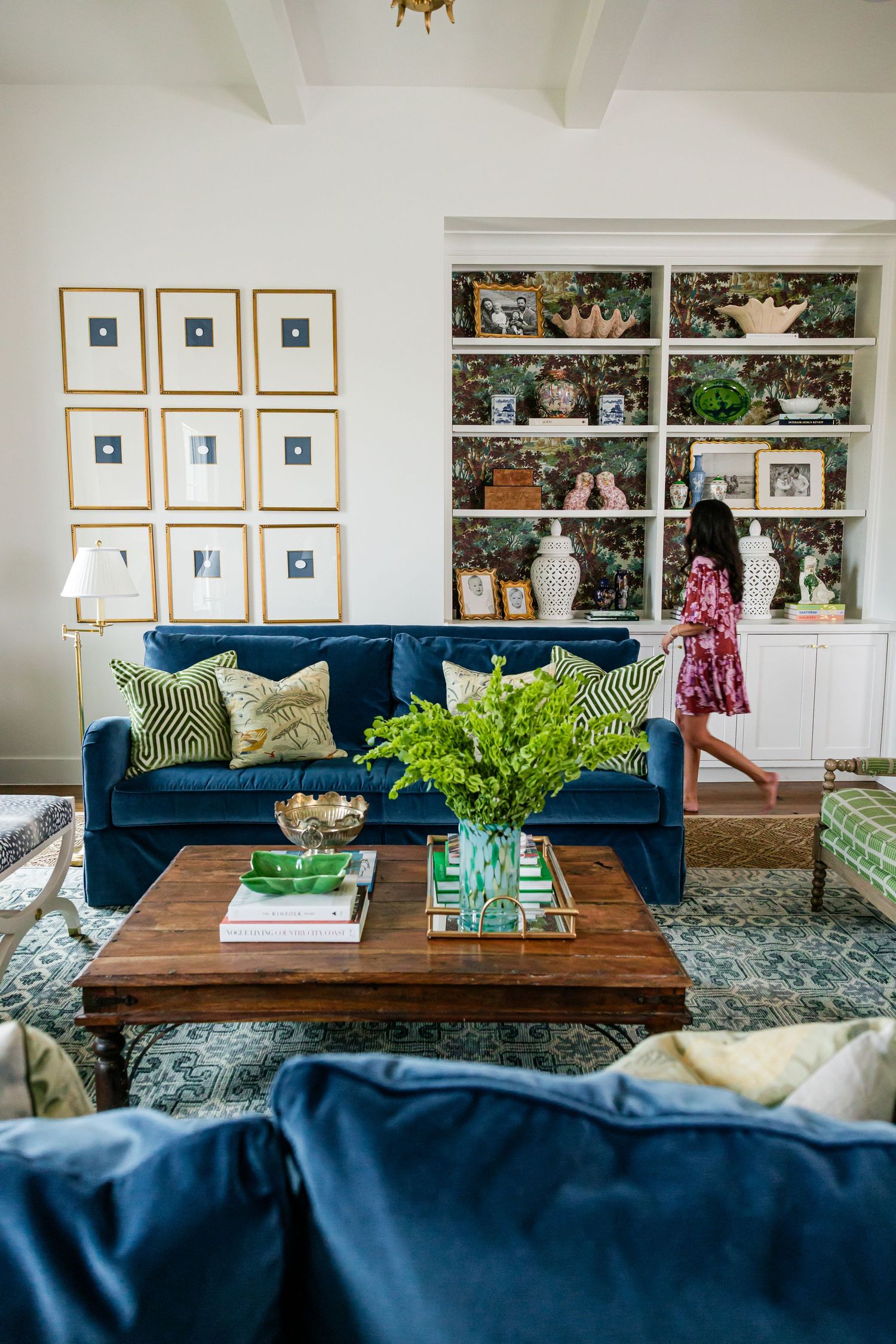 A bright living room features navy blue velvet sofas, green pillows, wood coffee table, and built-in white bookshelves.