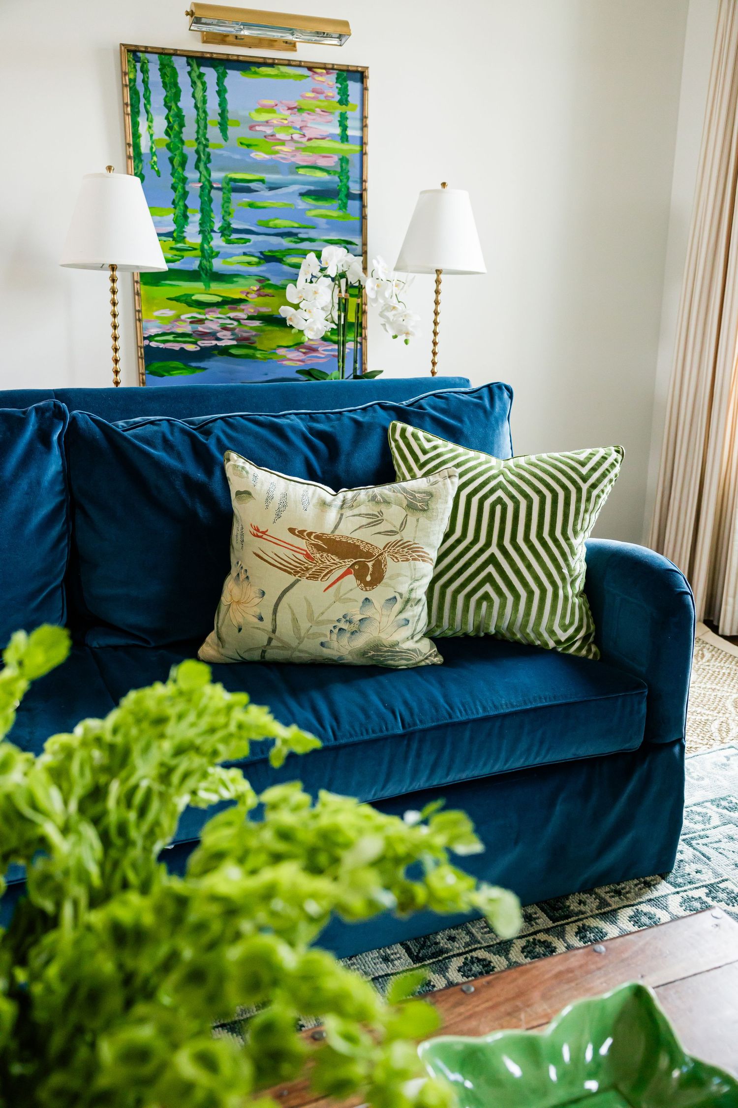 Blue velvet sofa with green and floral pillows in front of a nature-inspired painting.